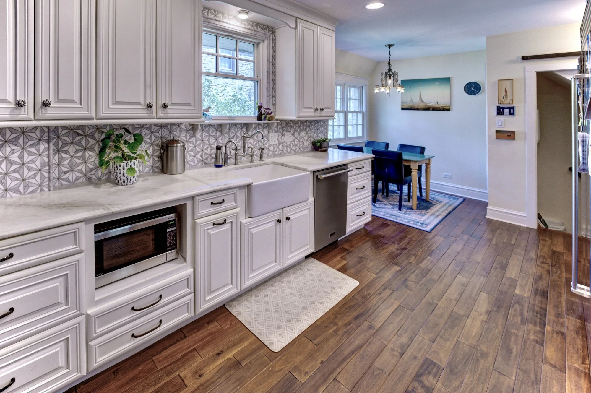 A bright kitchen with white cabinetry, a marble countertop, a farmhouse sink, and decorative patterned backsplash tiles. A potted plant sits on the counter, and a microwave is built into the lower cabinets. Adjacent to the kitchen is a dining area wi