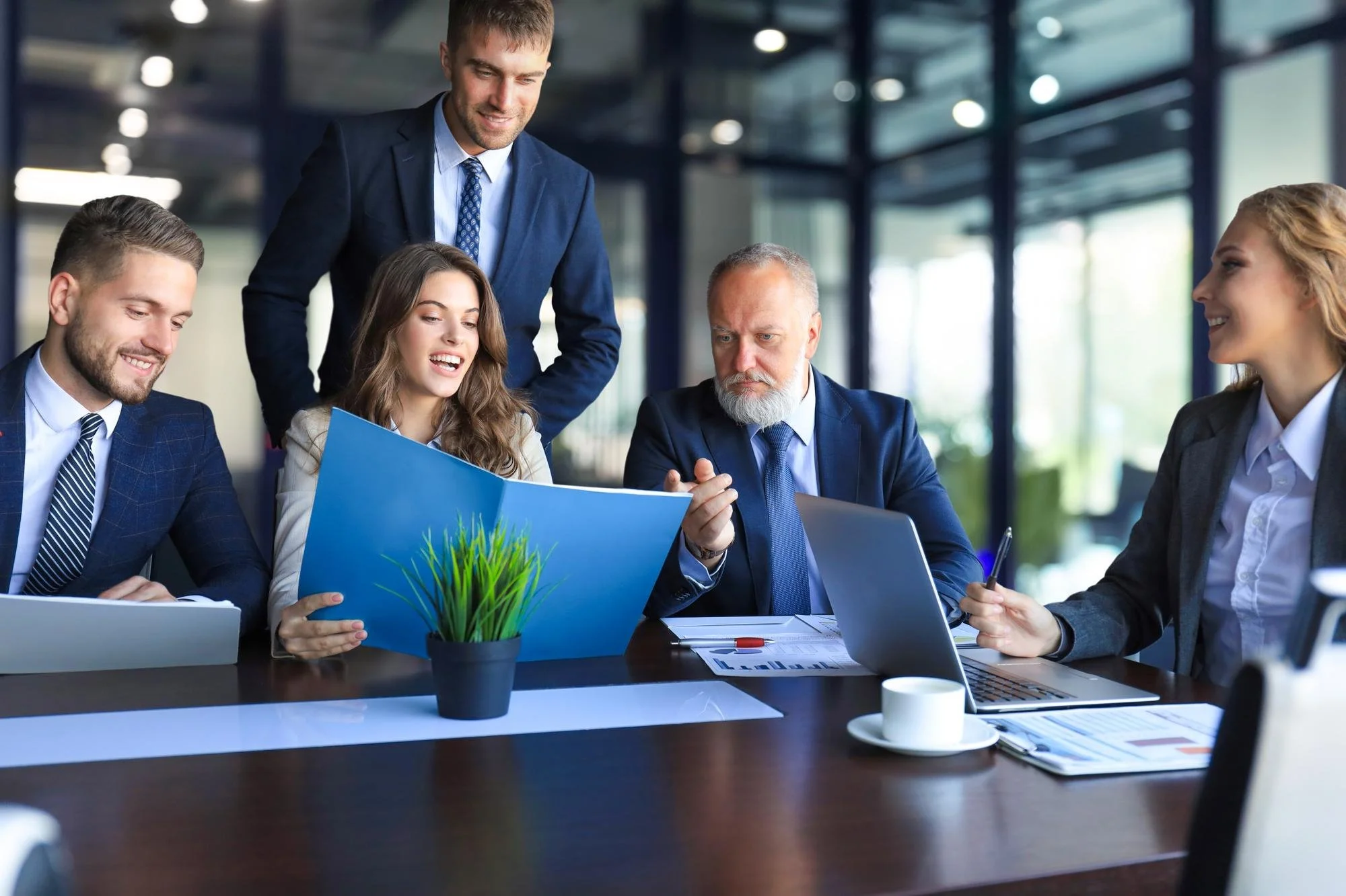 Group of five business professionals gathered around a table in a modern office, reviewing a folder with charts and graphs, with a small potted plant, coffee cups, and laptops on the table.