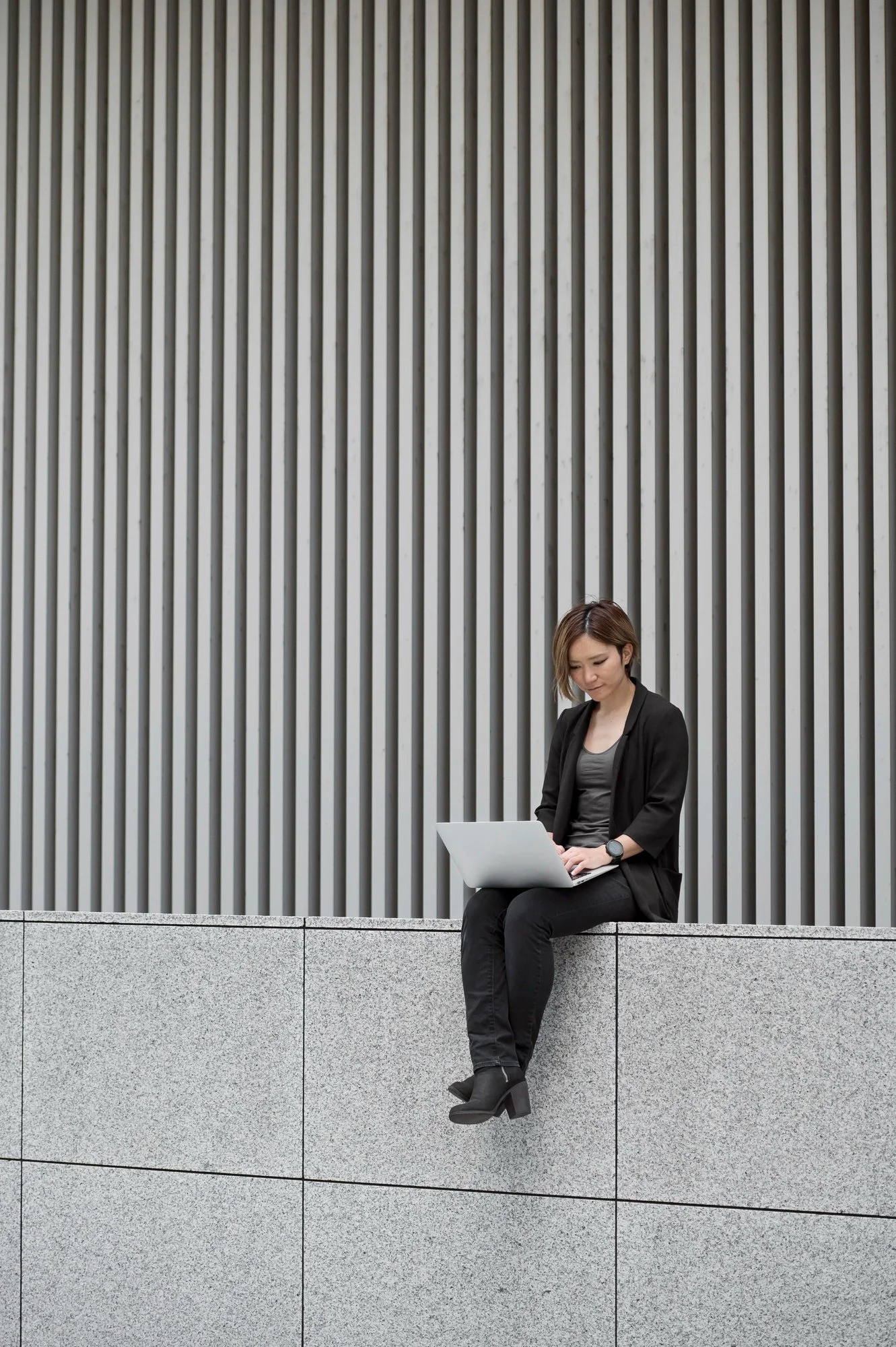 A young woman sitting on a concrete ledge in front of a modern building wall, working on a laptop. She has short hair, is dressed in black, and wears a watch and chunky black heeled boots.