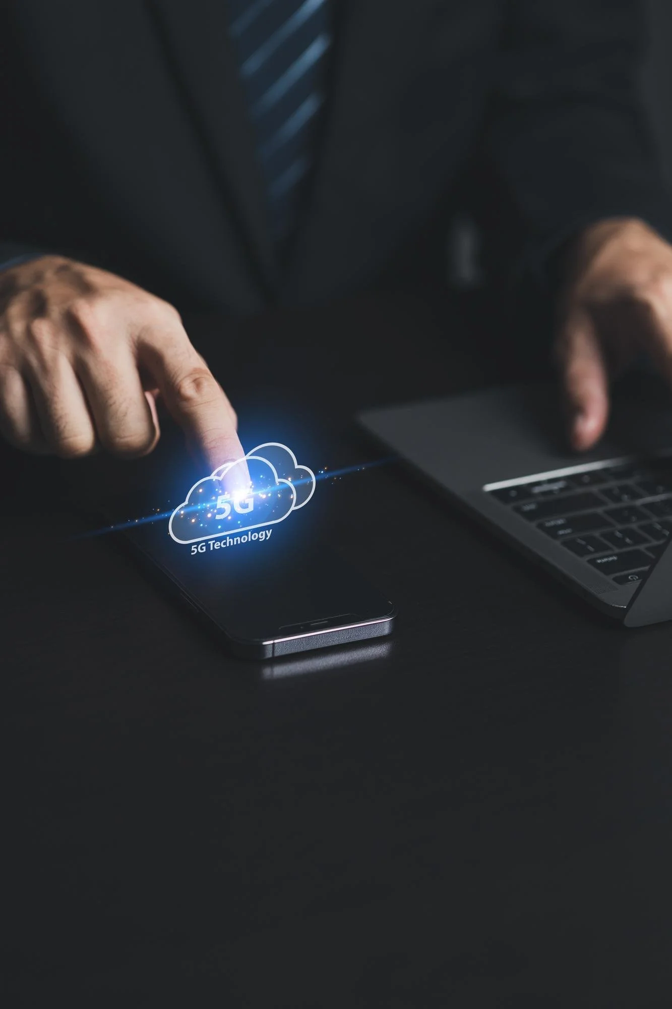 A person in a suit using a smartphone with a holographic display of a cloud icon labeled '5G Technology' and a laptop nearby.
