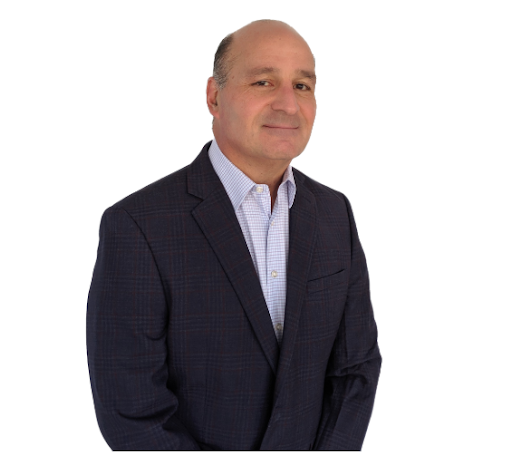 A middle-aged man wearing a dark plaid blazer and a light blue dress shirt, standing with a slight smile against a plain white background.
