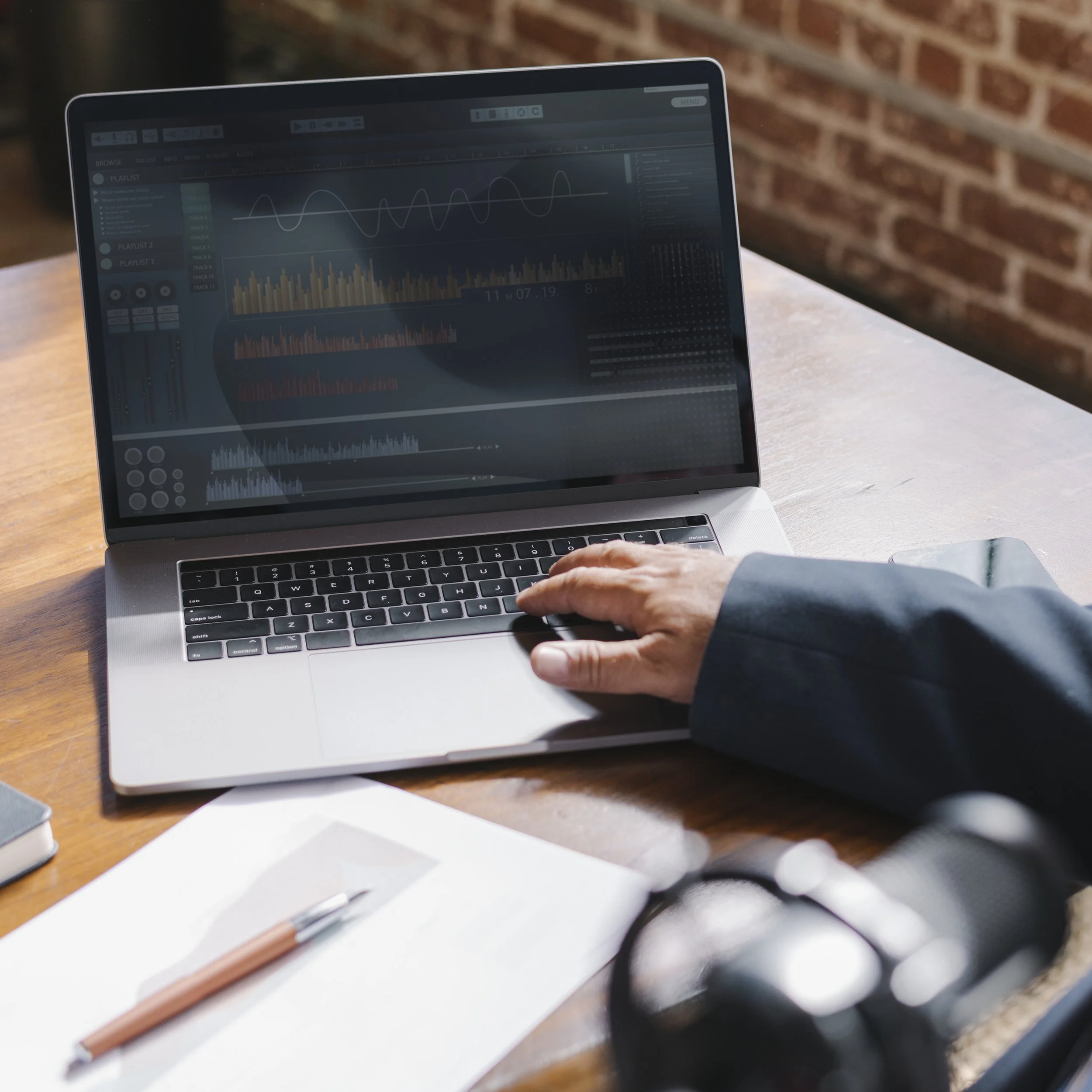 A person using a laptop on a wooden table, with a brick wall in the background, displaying audio editing software with waveforms and sound levels.
