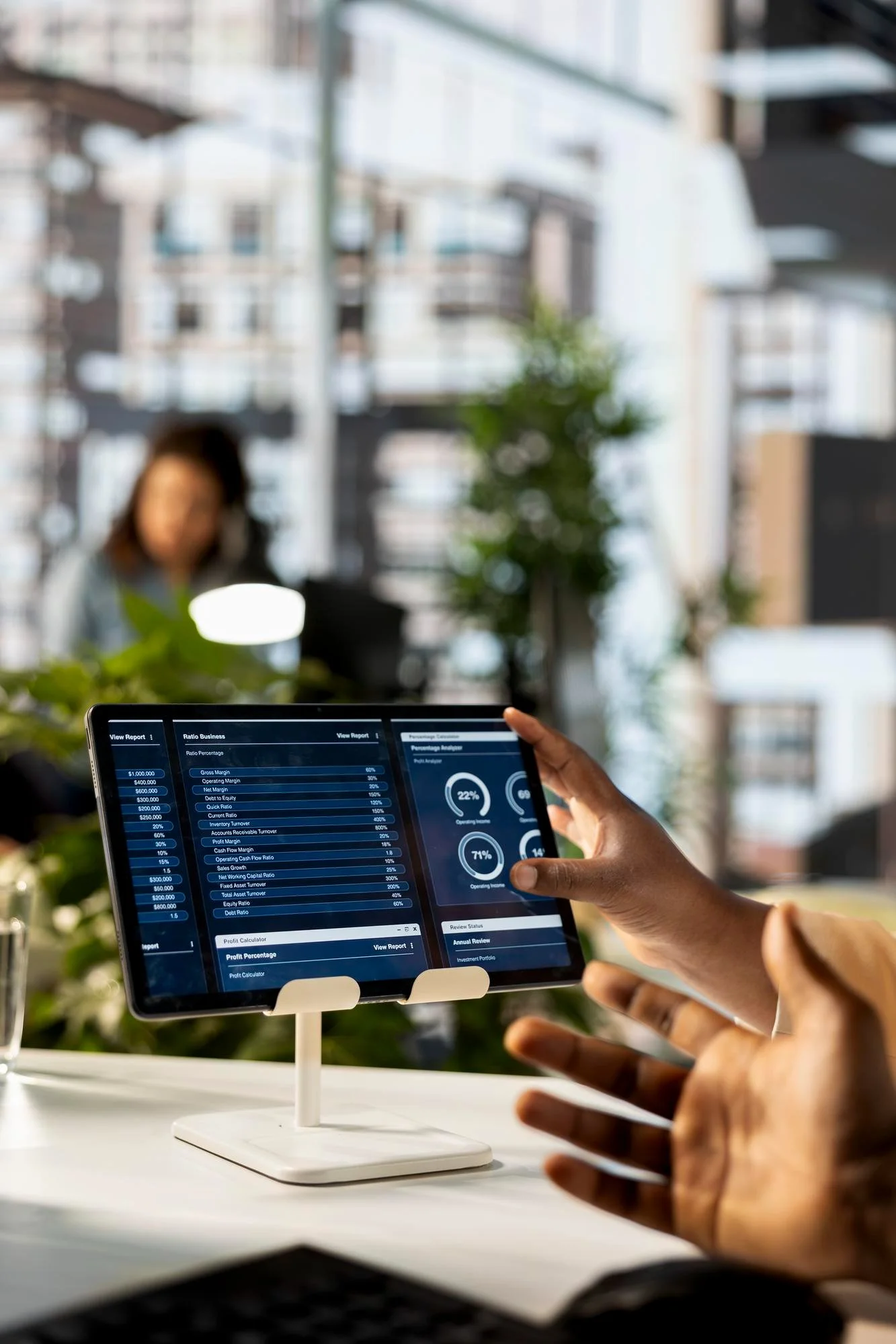 A person holding a tablet displaying financial data and charts, with the tablet mounted on a stand on a white desk, in a modern office with large windows and a woman working in the background.