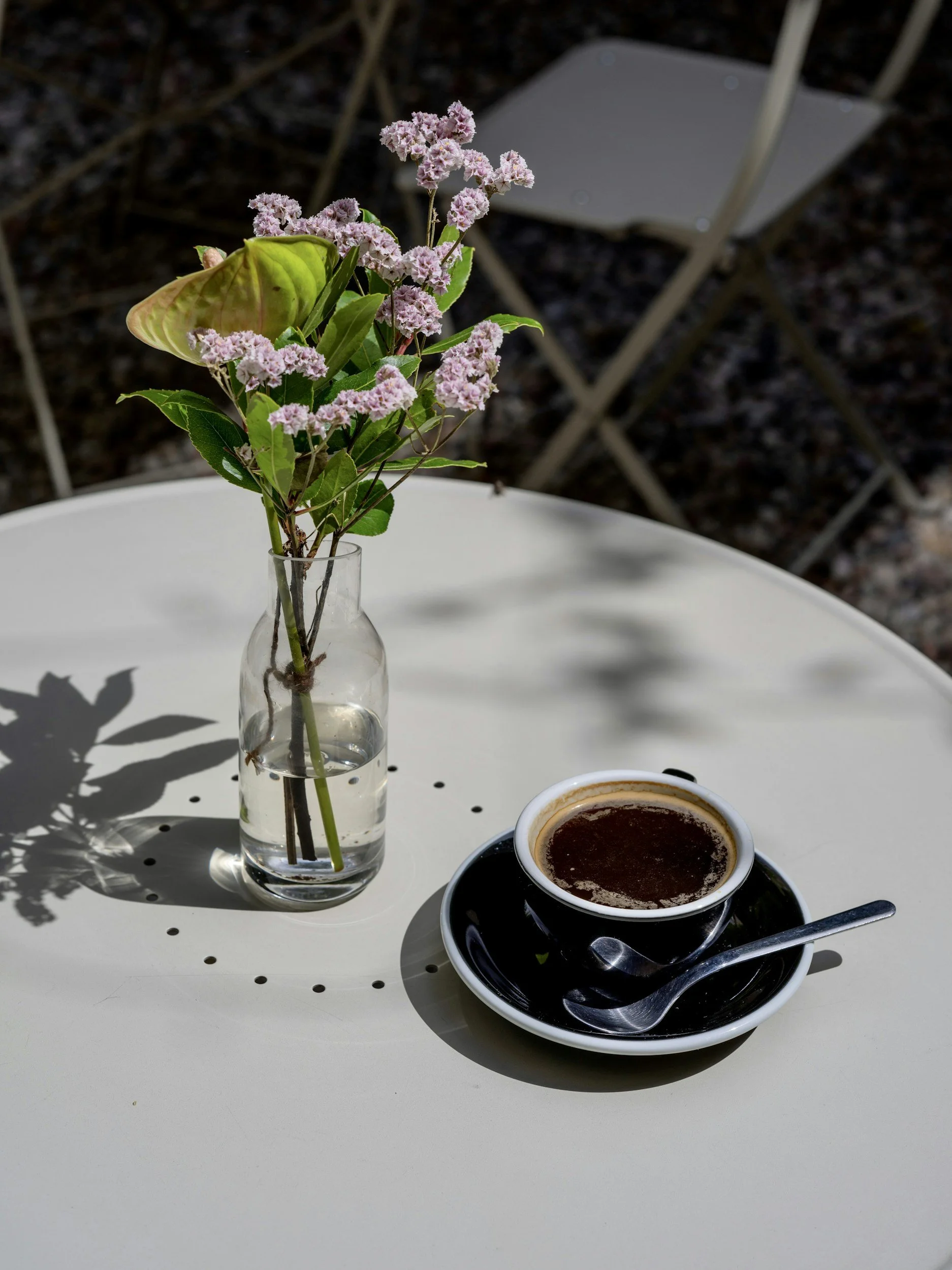 A small glass vase with pink flowers and green leaves on a white table, with a cup of coffee and a spoon next to it, in an outdoor setting.