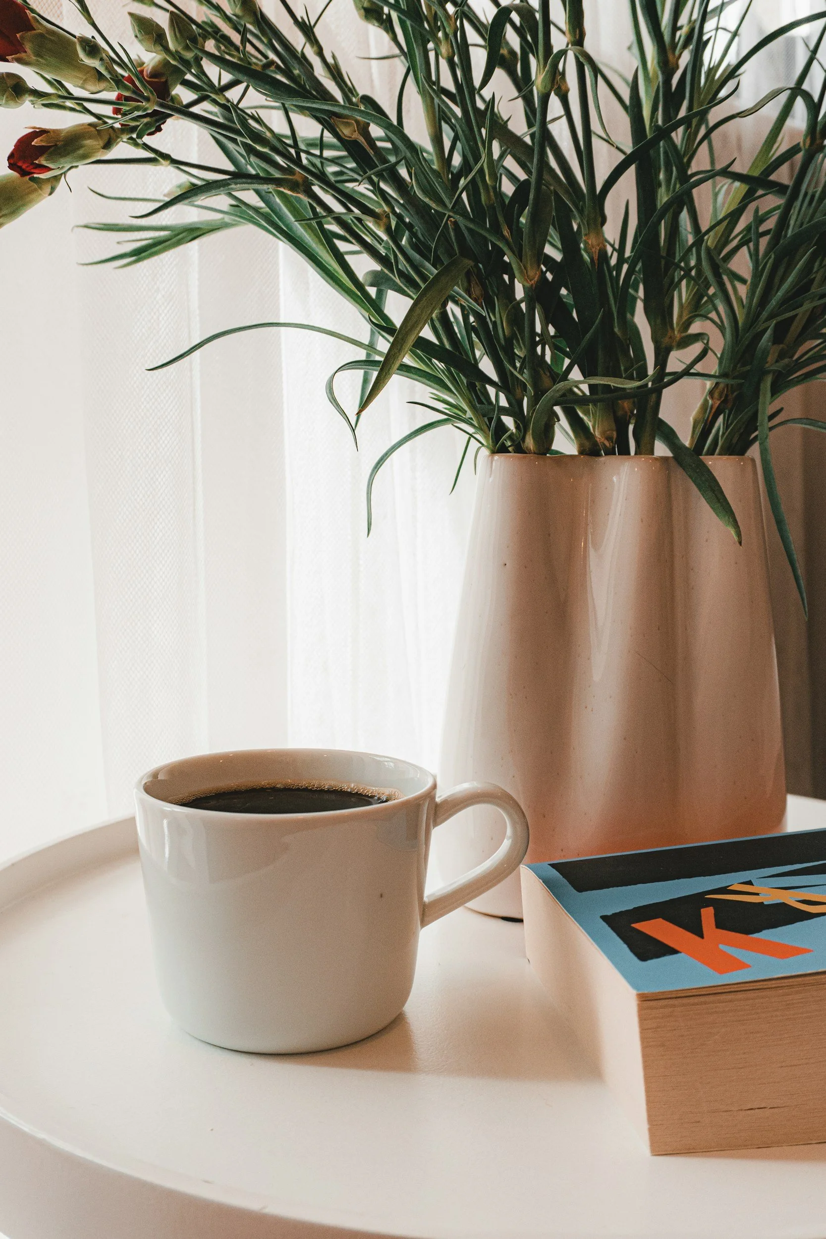 Coffee cup, books, and a vase of greenery on a sunlit side table