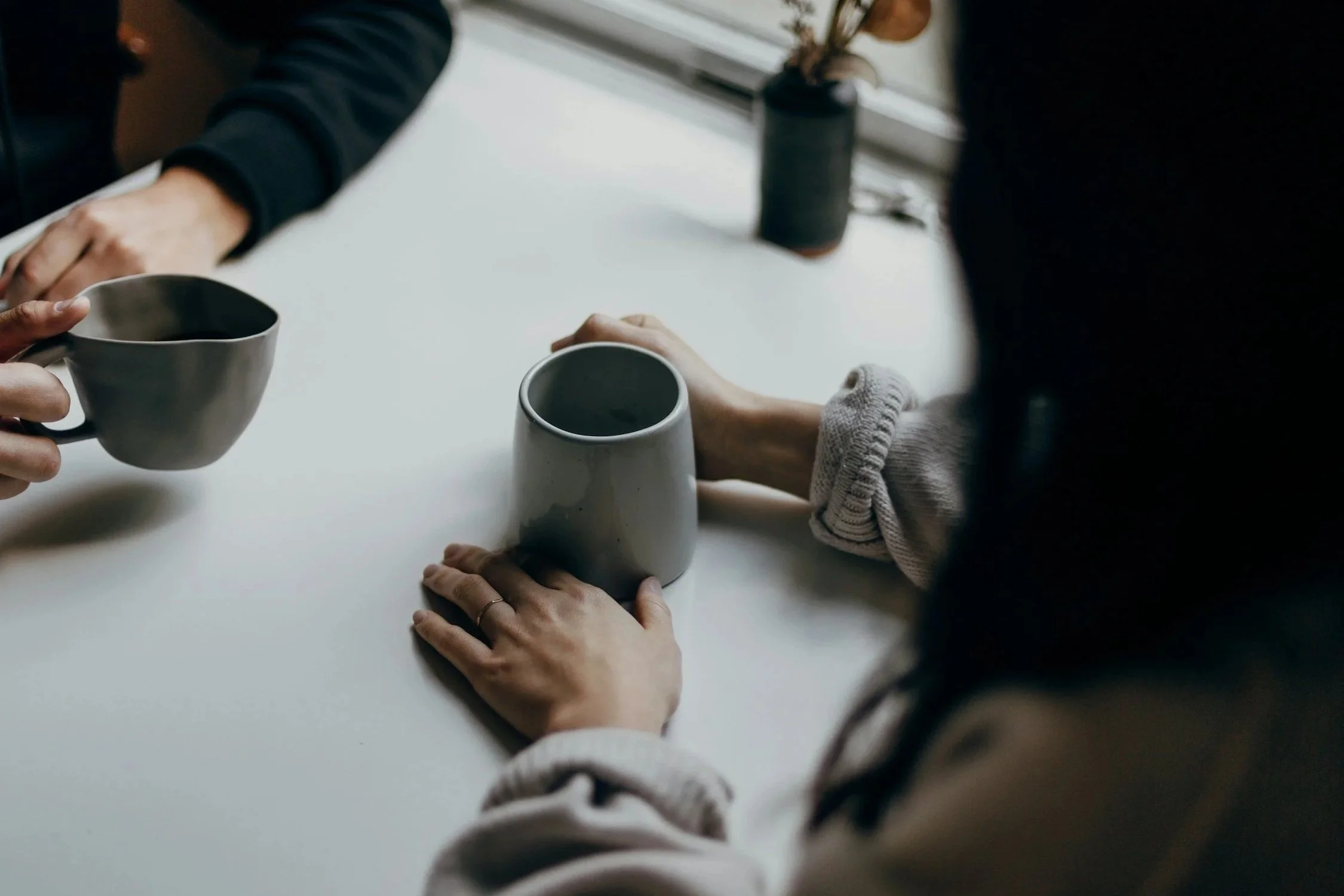 Two people sitting at a table holding warm mugs, in conversation.