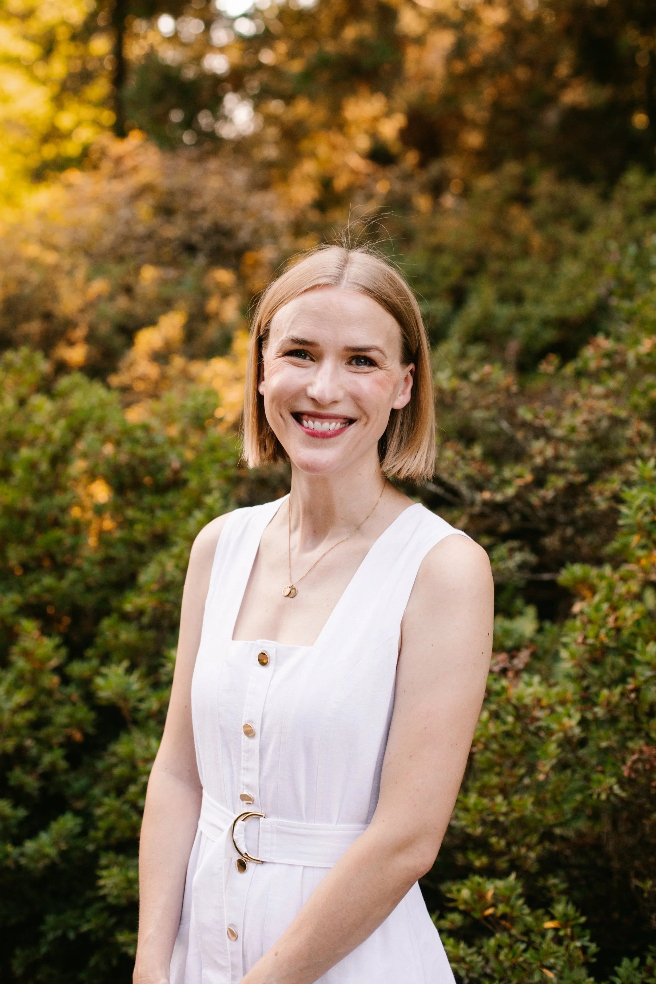 Therapist smiling outdoors in front of autumn foliage. She has blonde hair and is wearing a white sleeveless dress with gold buttons.