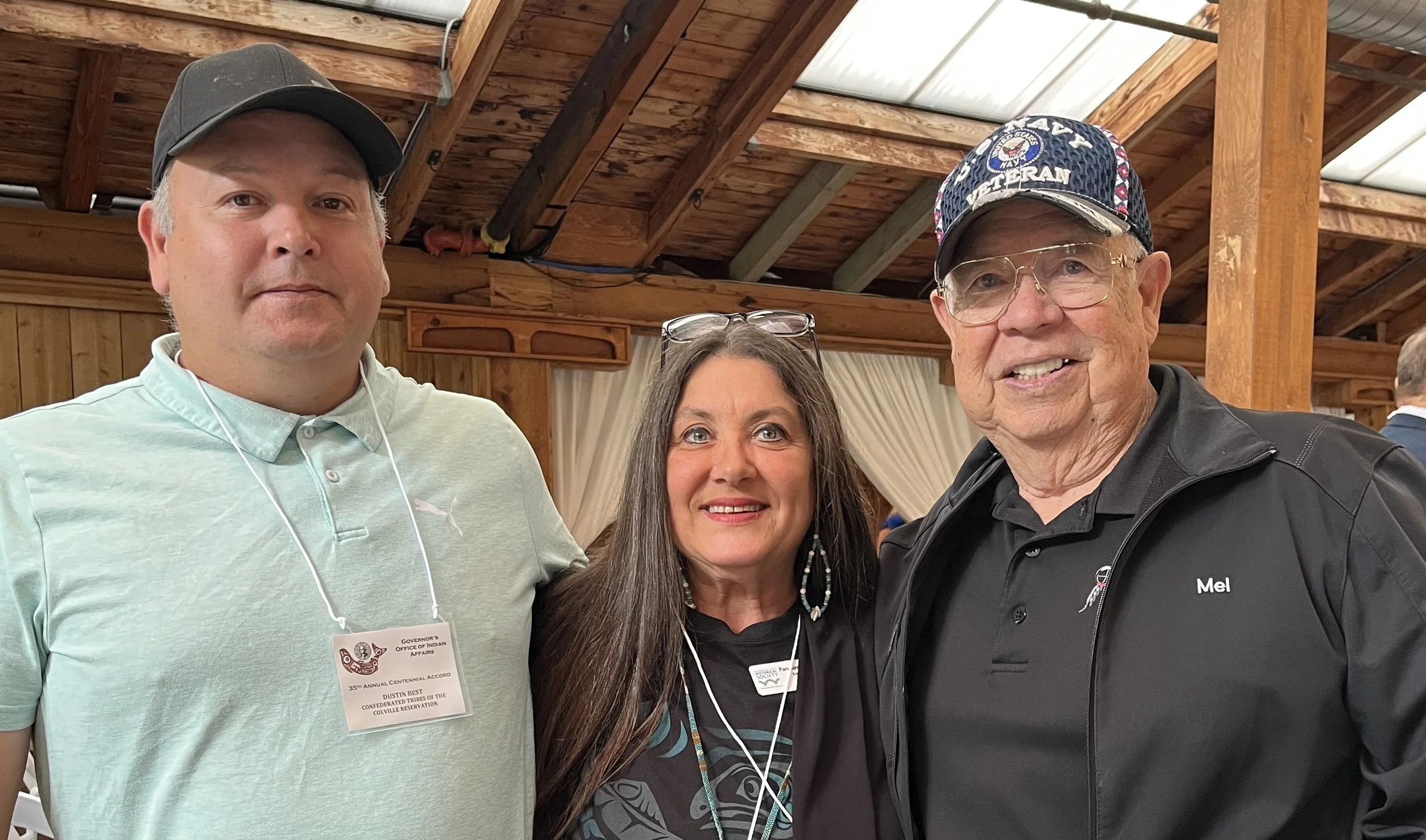 Three people taking a group photo at an indoor event with wooden ceiling beams, dressed casually, with two men and a woman smiling at the camera.
