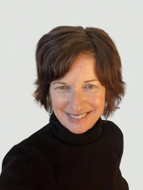 A woman with short brown hair wearing a black turtleneck, smiling at the camera against a plain background.