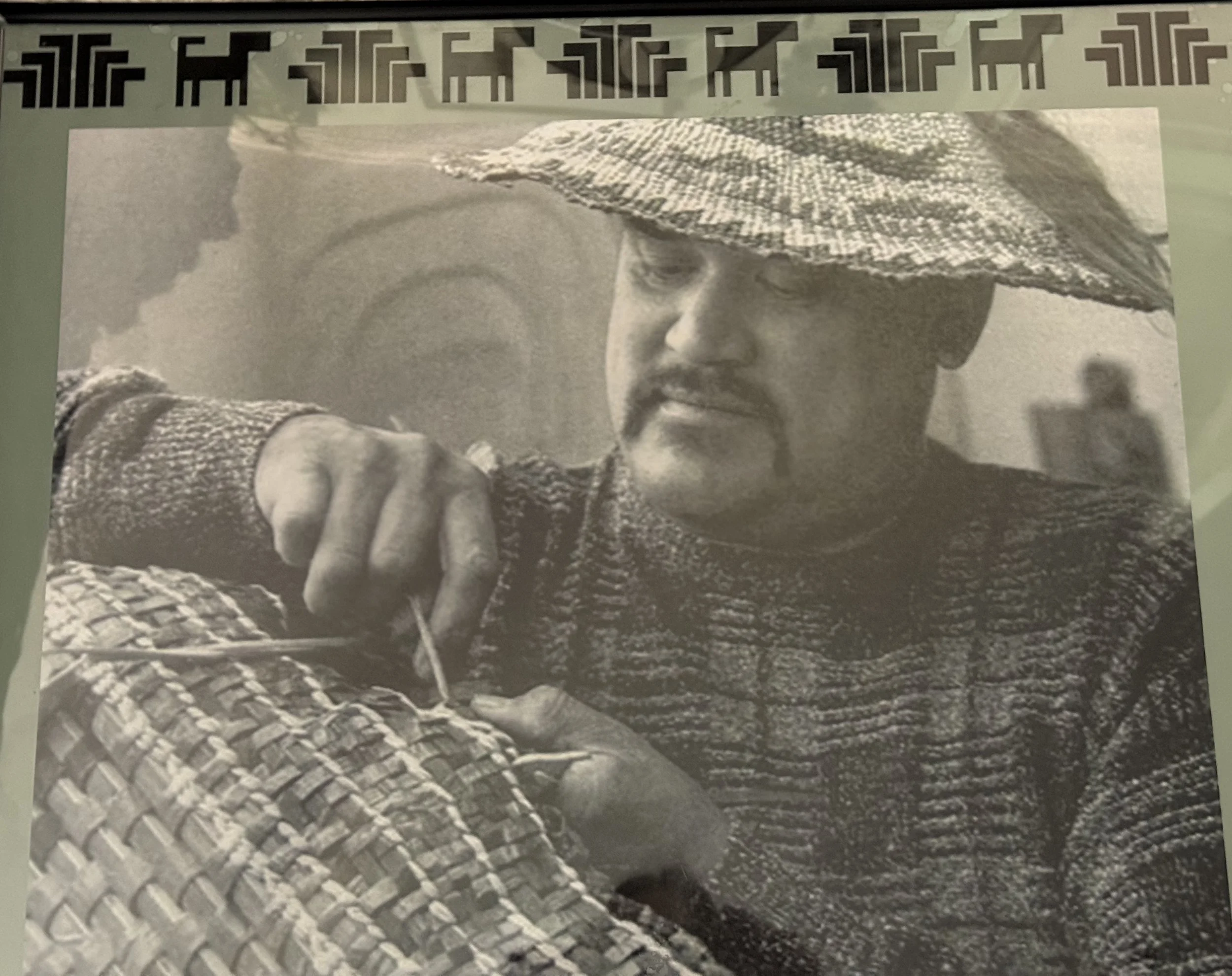 Black and white photo of a man with a beard and moustache wearing a wide straw hat, pointing towards the camera with a serious expression. The background is blurred with a hint of other figures.