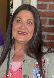 A woman with long dark hair smiling, standing in front of a brick wall, wearing a light-colored jacket with floral patterns and a pink top.