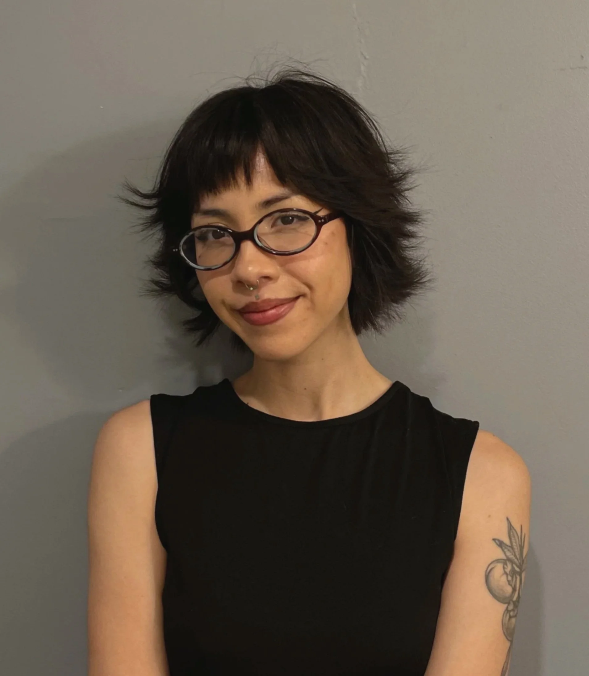 Close-up photo of a woman with short, layered dark hair wearing glasses and a black sleeveless top. She has a septum piercing and a tattoo of leaves on her right arm, against a gray wall background.