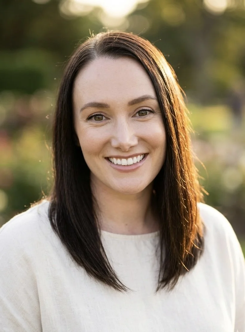 A woman with dark brown hair and light skin smiling outdoors during golden hour with sunlight on her hair, wearing a white top.