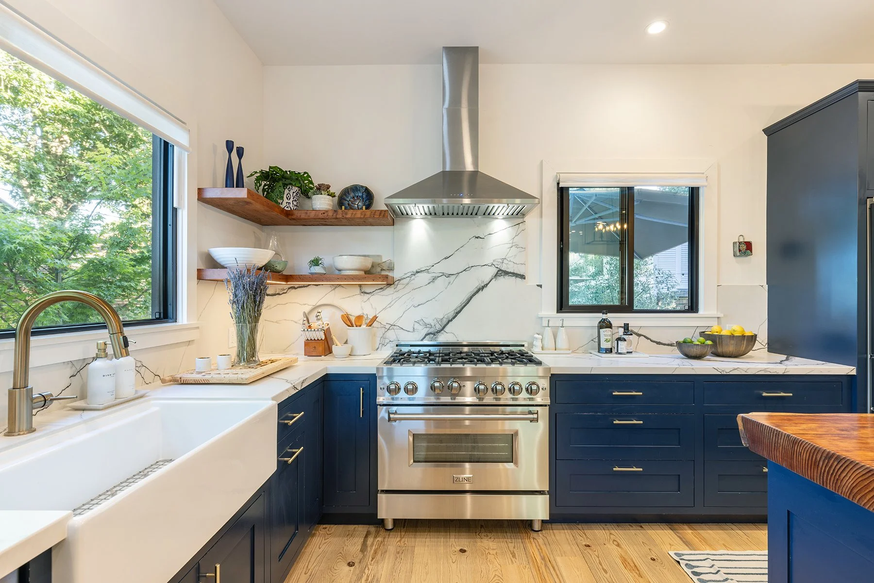 Kitchen with blue cabinetry, stainless steel stove, white marble backsplash, wooden open shelves with decorative items, large window showing green trees, and a white farmhouse sink.