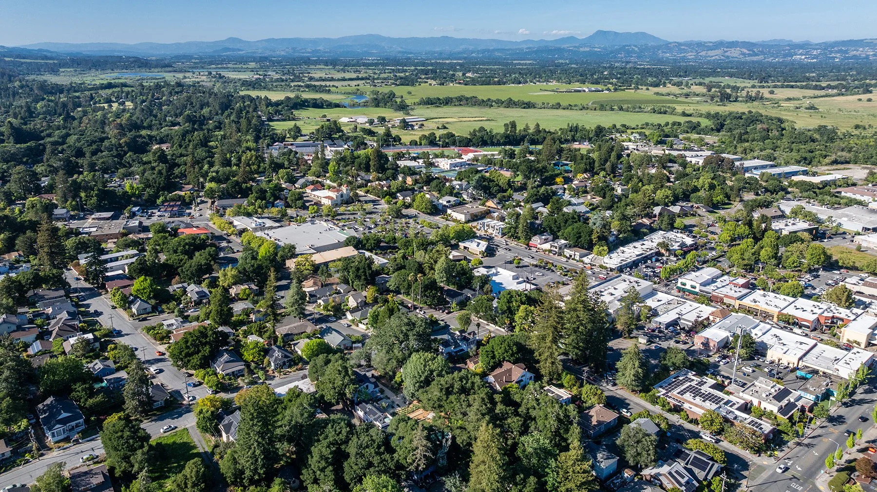 An aerial view of a small town with residential houses, commercial buildings, and trees, set against a backdrop of green fields and distant mountains under a blue sky.