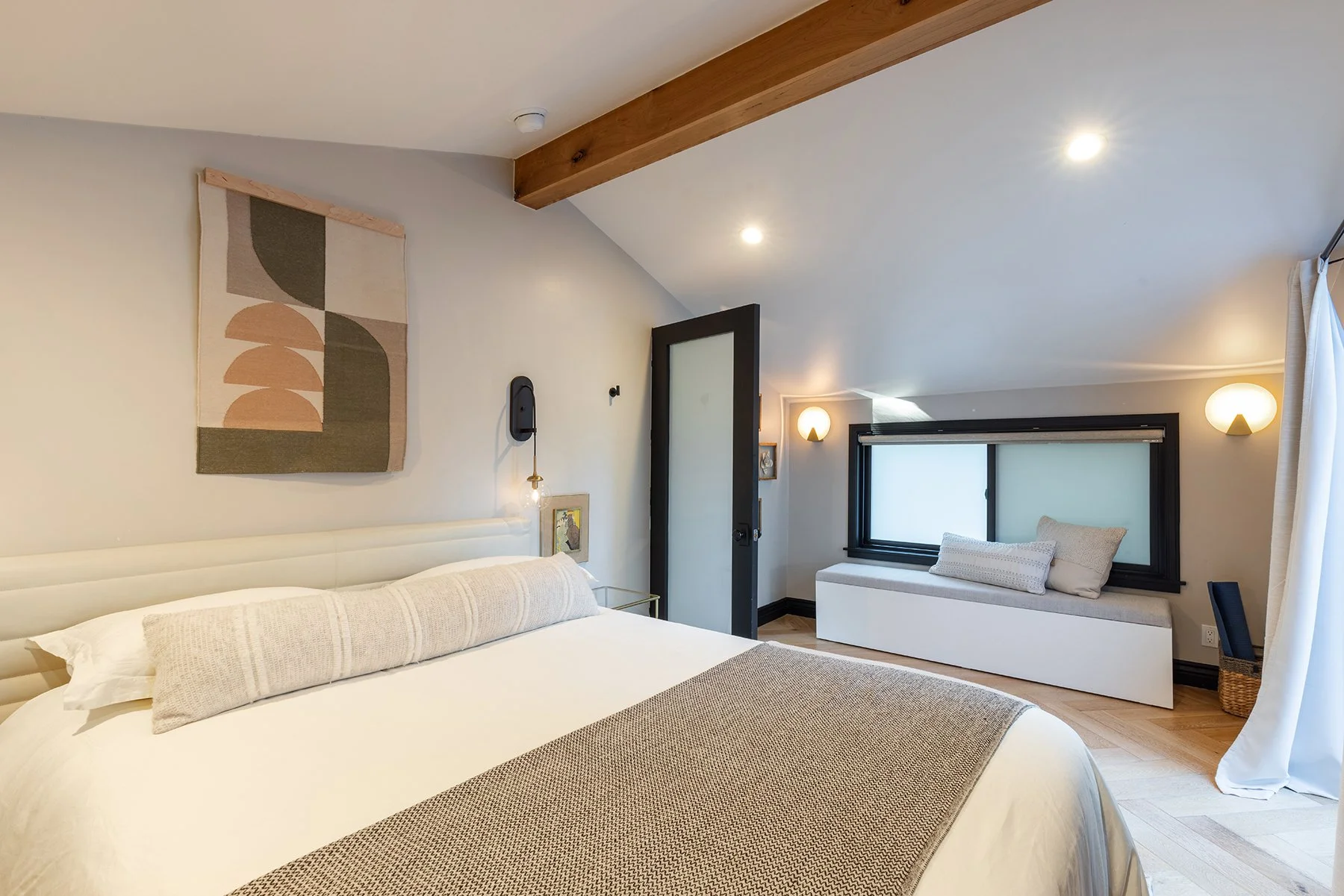 A cozy bedroom with a bed, decorative wall art, a window seat with pillows, and modern lighting.