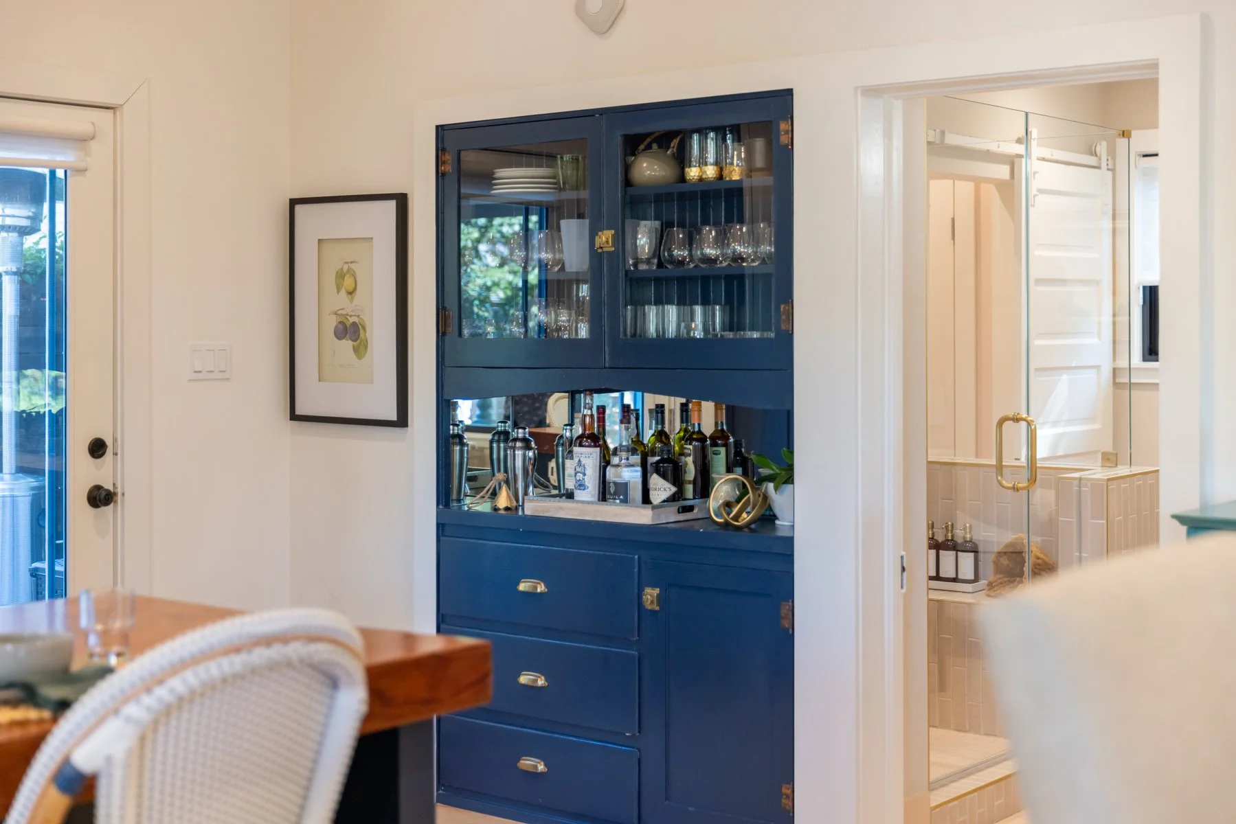 Blue kitchen cabinet with glass doors displaying glassware, bottles of alcohol, and bar accessories; adjacent to a bathroom with a glass shower door.