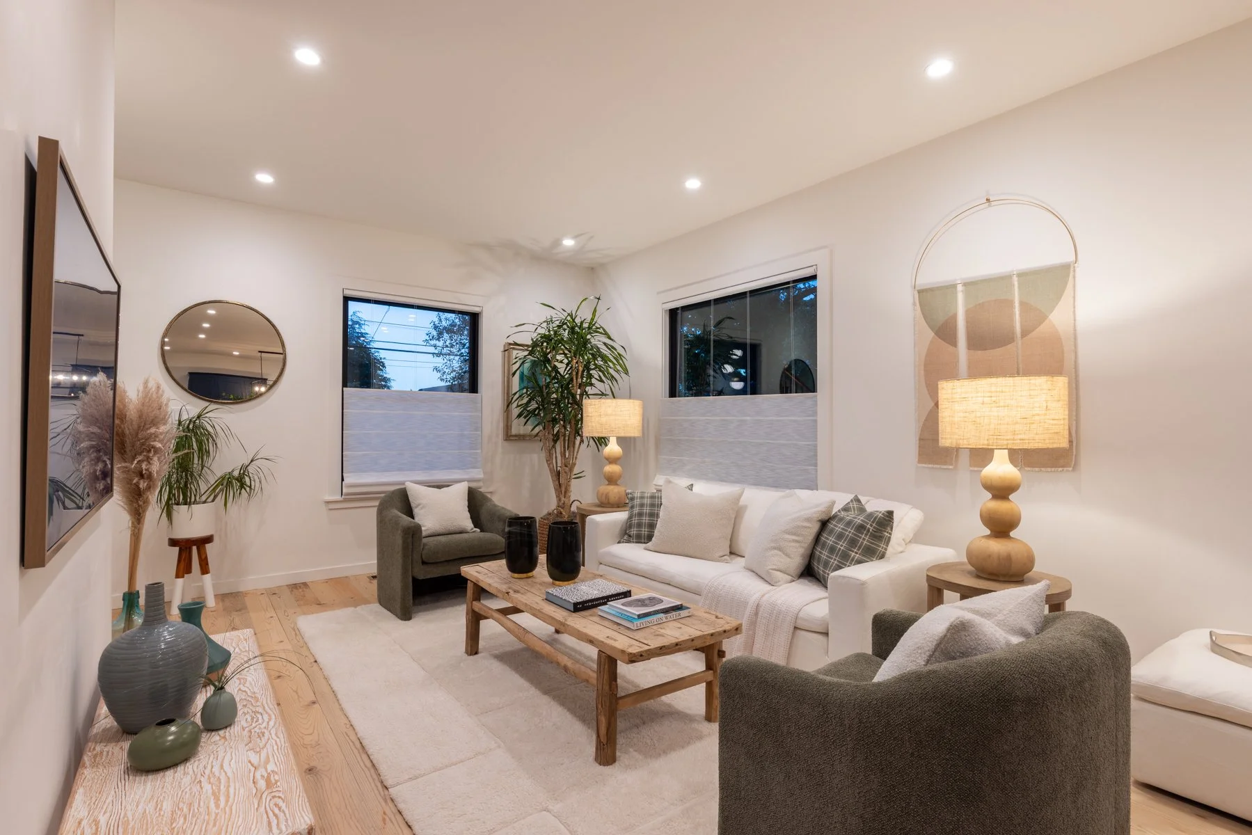 Living room with white walls, wooden floor, two large windows, a white sofa with pillows, two gray armchairs, a wooden coffee table, potted plants, lamps, wall art, and decorative vases.