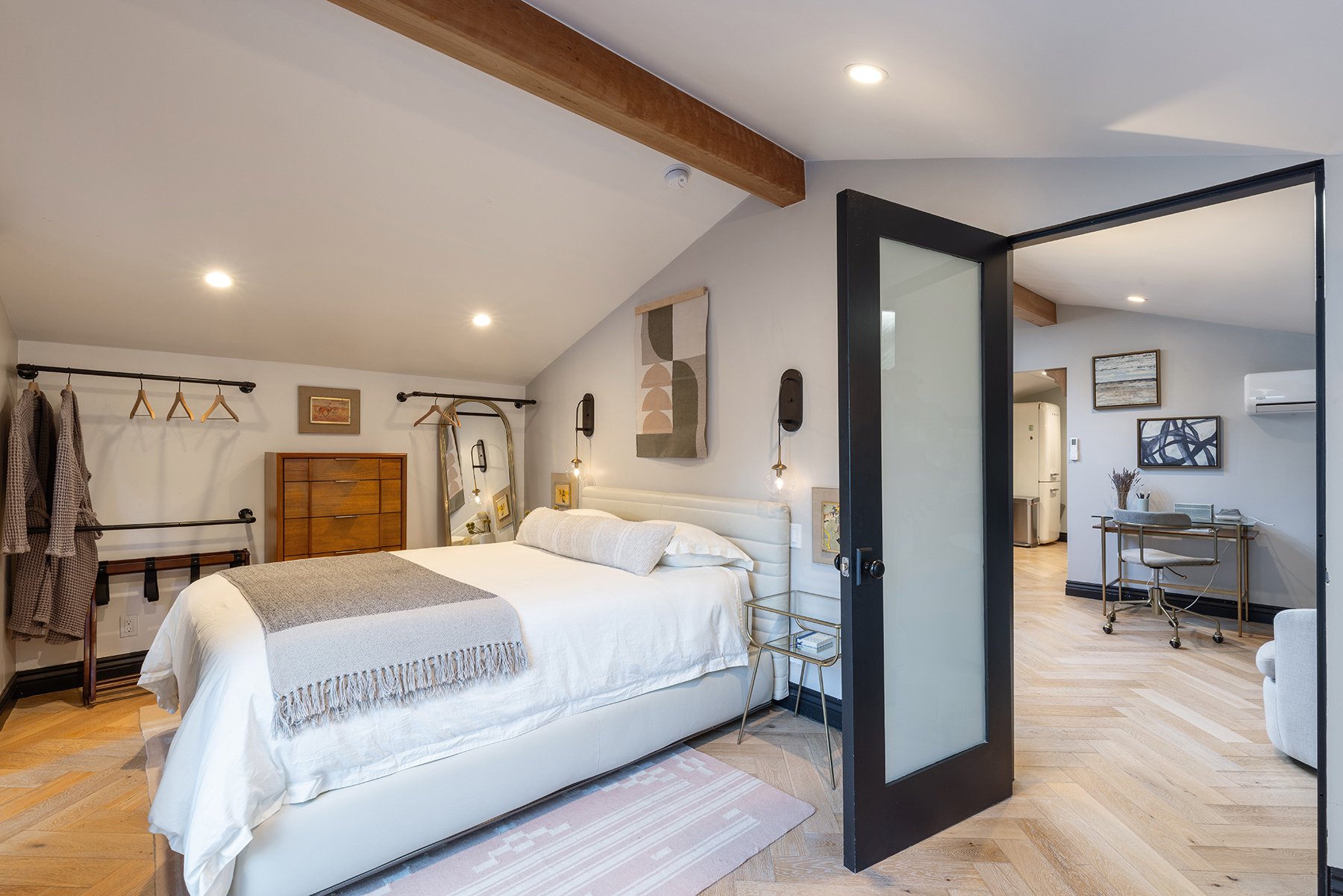 A modern bedroom with a white bed, gray and white decor, wall art, and a black sliding door leading to another room with a desk and chairs. There are wooden accents, hanging clothes, and warm lighting.