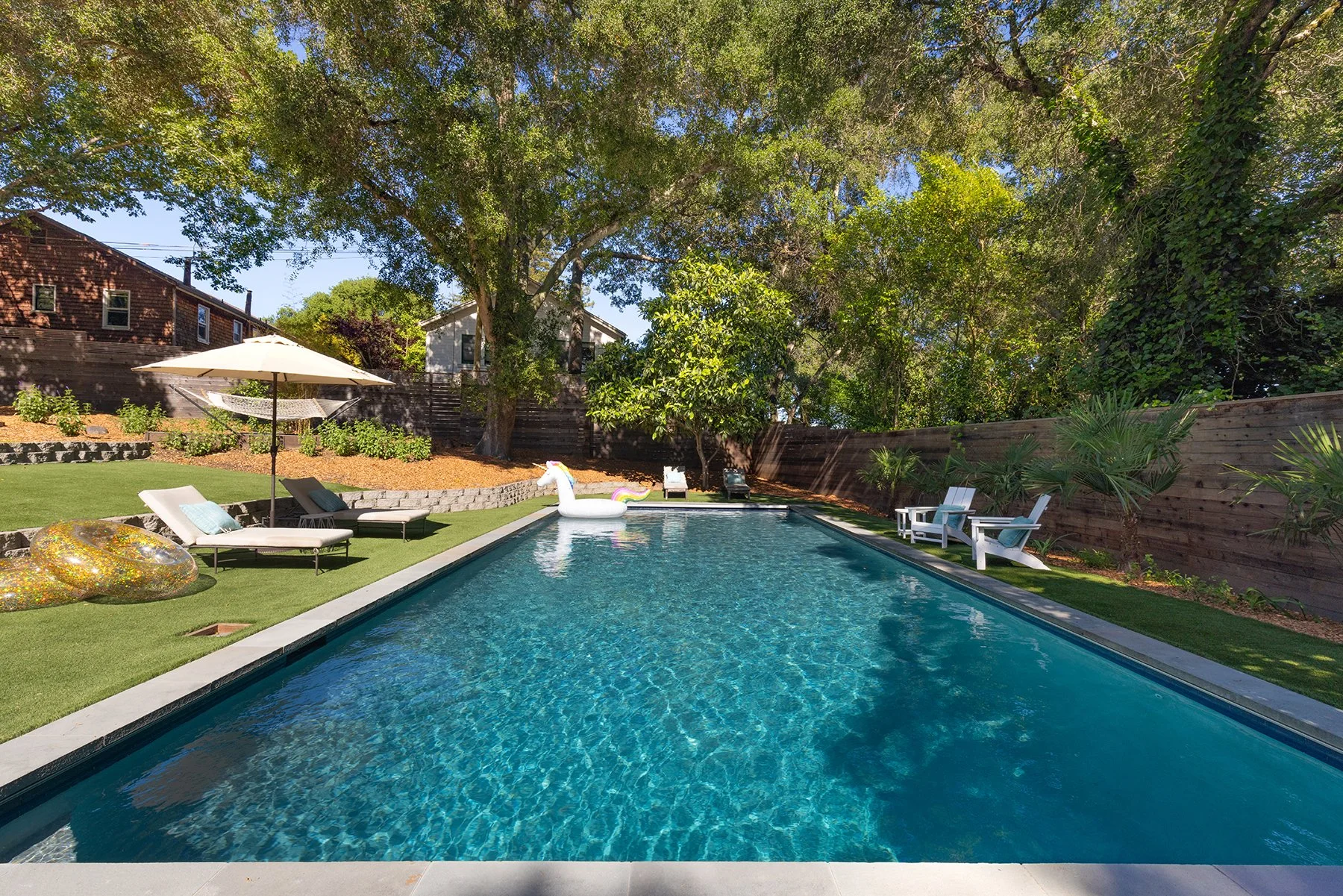 A backyard swimming pool surrounded by lounge chairs, umbrellas, and decorative inflatables, with large trees and a wooden fence in the background.