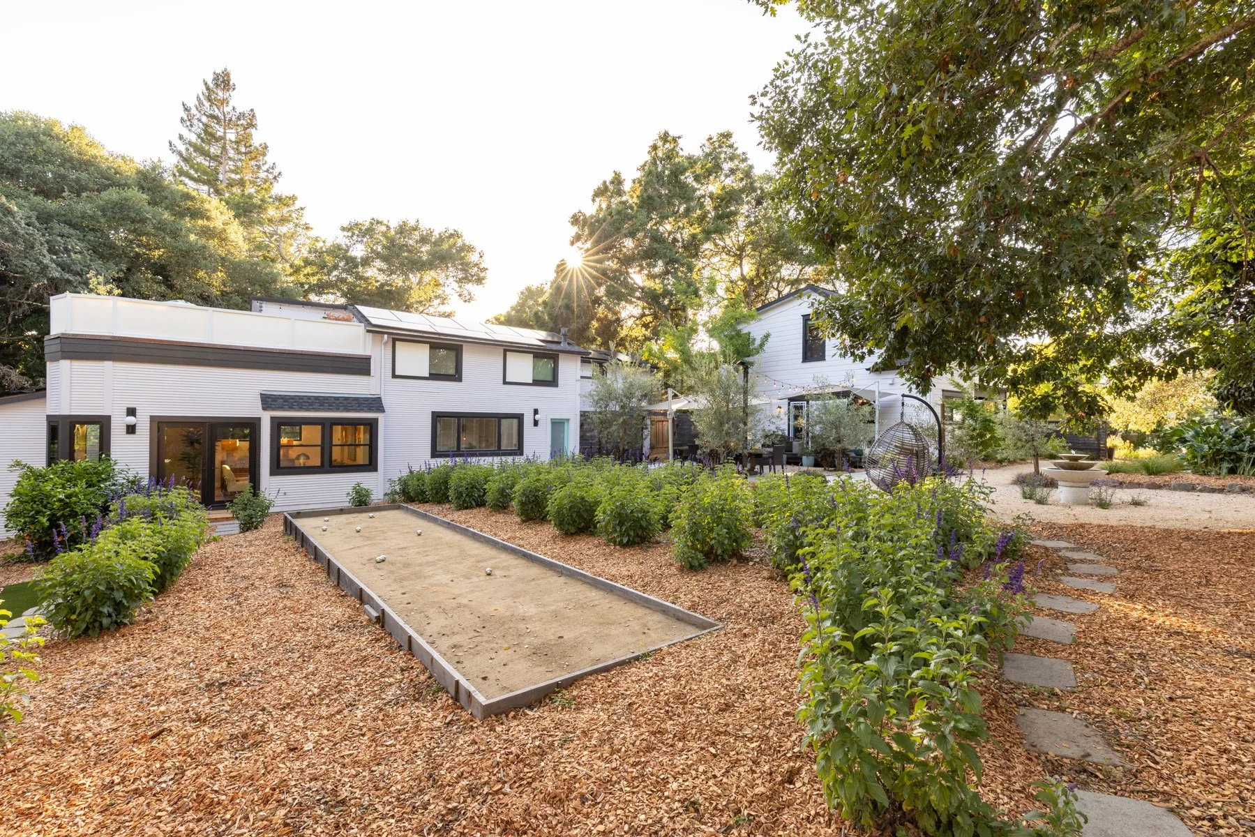 A backyard with a landscaped garden, a bocce ball court, and two modern white houses with black trim, surrounded by tall trees and lush greenery, during sunset.