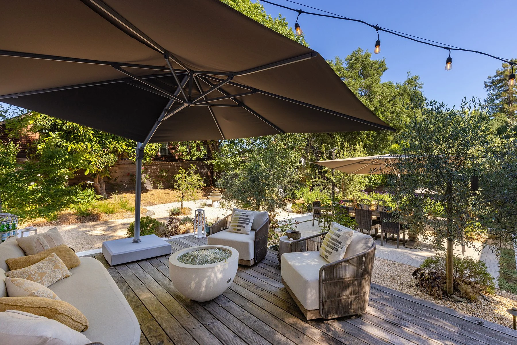 Outdoor patio with seating area, large umbrella, string lights, trees, and a landscaped garden.