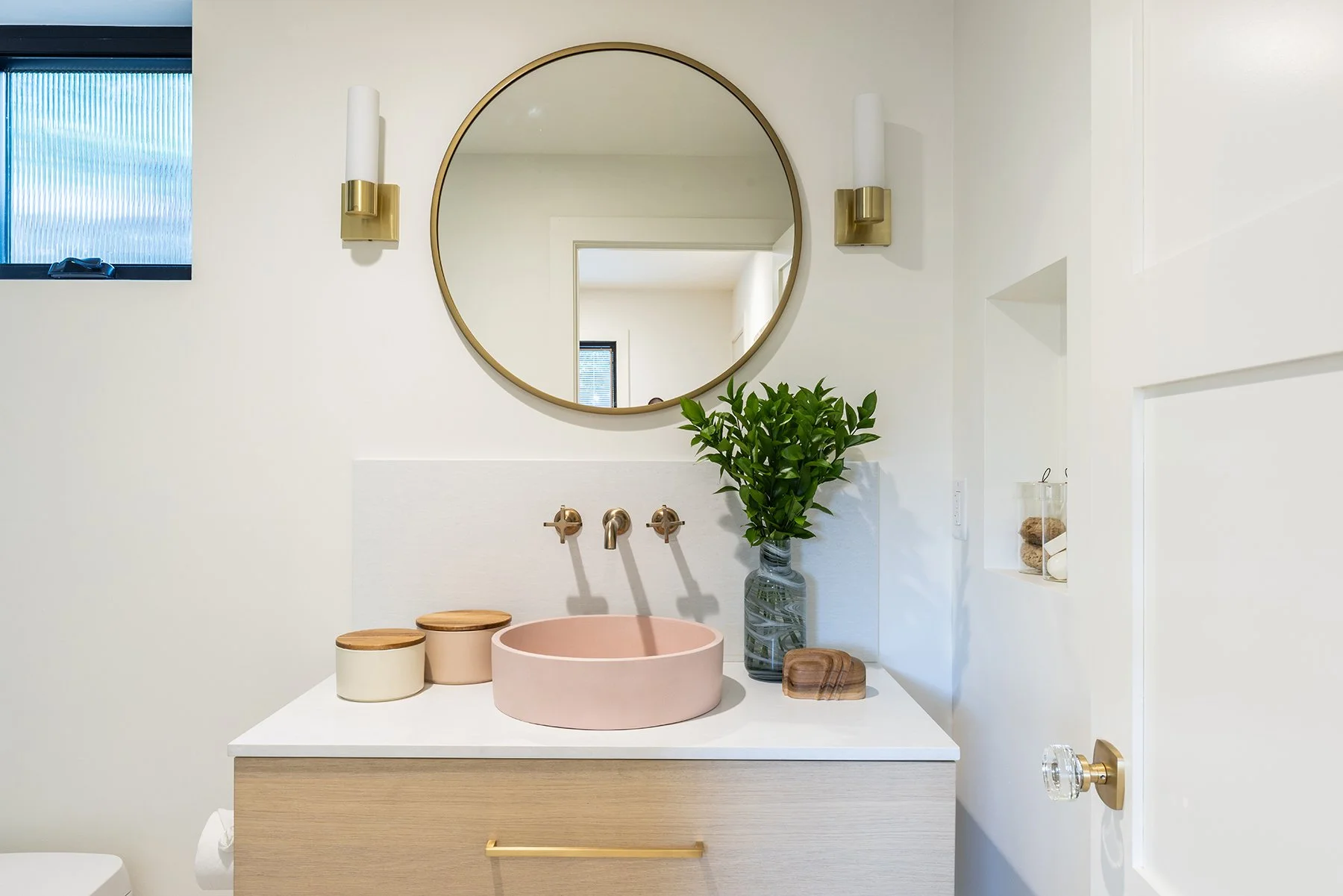 A modern bathroom vanity with a round mirror, wall-mounted brass light fixtures, a pink vessel sink, and greenery in a glass vase. There are small containers and wood accents on the white countertop, with a window and light-colored walls.