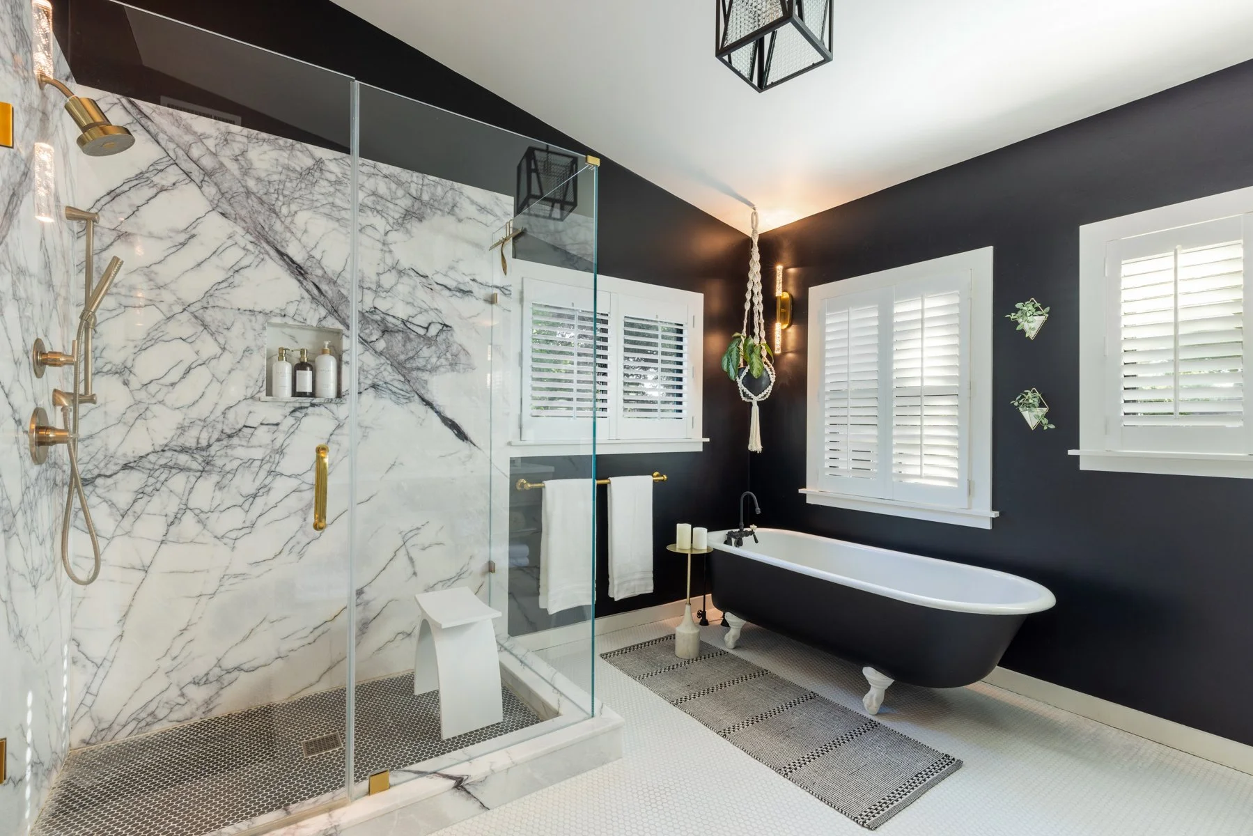 Modern bathroom with black and white color scheme, featuring a walk-in shower with marble walls and gold fixtures, a clawfoot bathtub near two windows with white shutters, and green plants hanging on the wall.