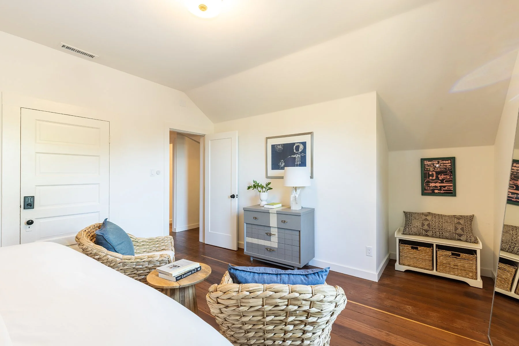 Living room with white walls, hardwood floors, and minimalistic decor including a gray dresser with a white lamp and green plant, wicker chairs with blue cushions, and artwork on the walls.