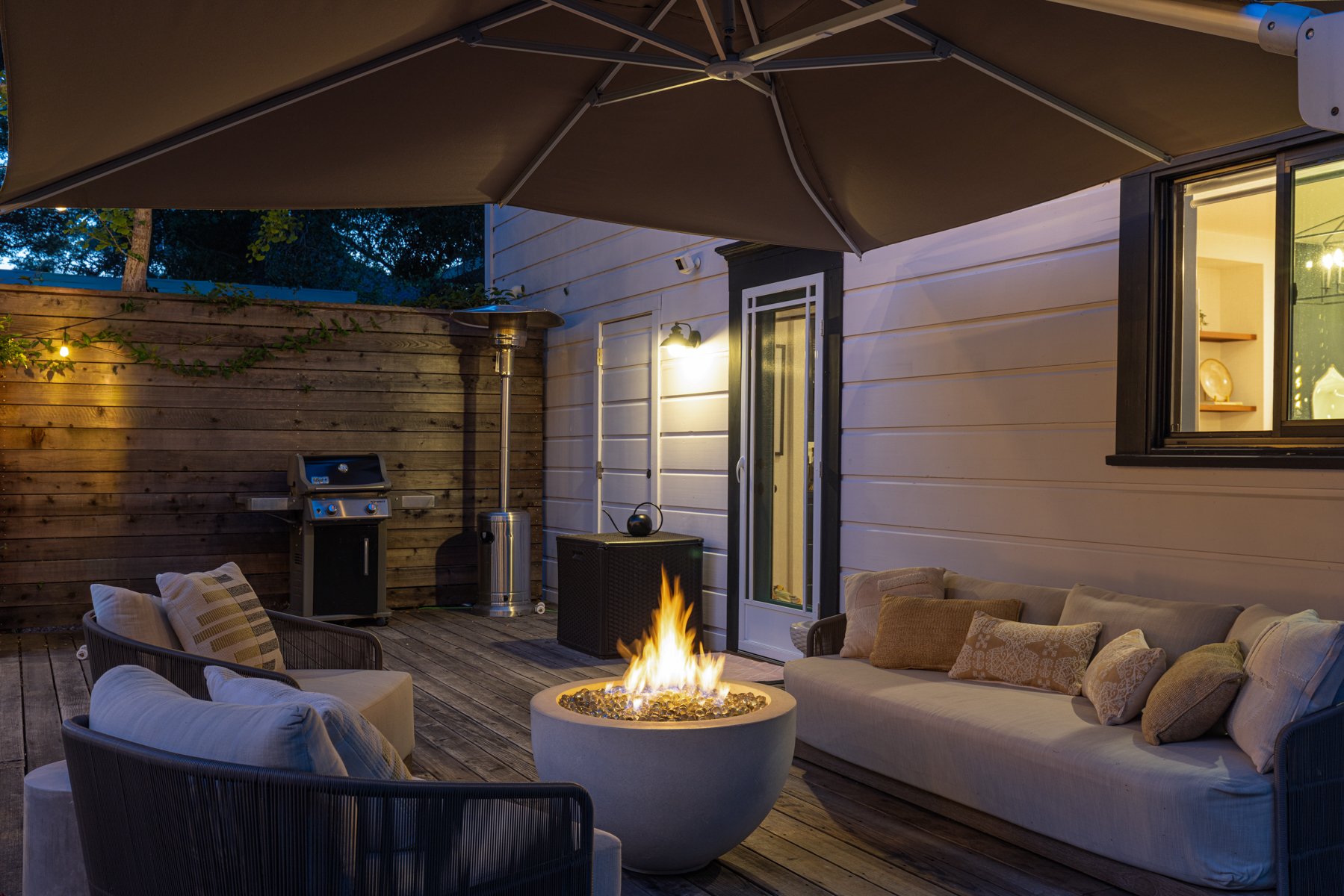 Outdoor patio at dusk showing a fire pit, cushioned seating, an umbrella, a grill, and a heater against a wooden fence, with string lights and house lighting.