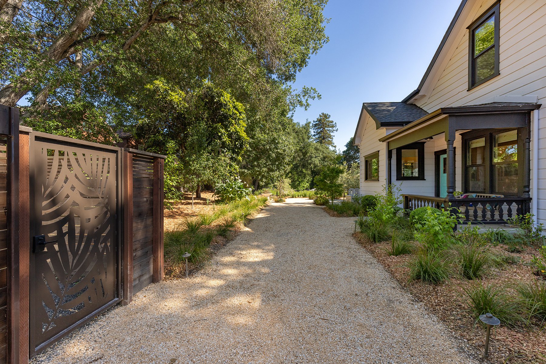 A gravel pathway next to a white house with black window trims and a small porch. Green shrubs and plants line the sides of the path, and large trees are seen in the background under a clear blue sky.