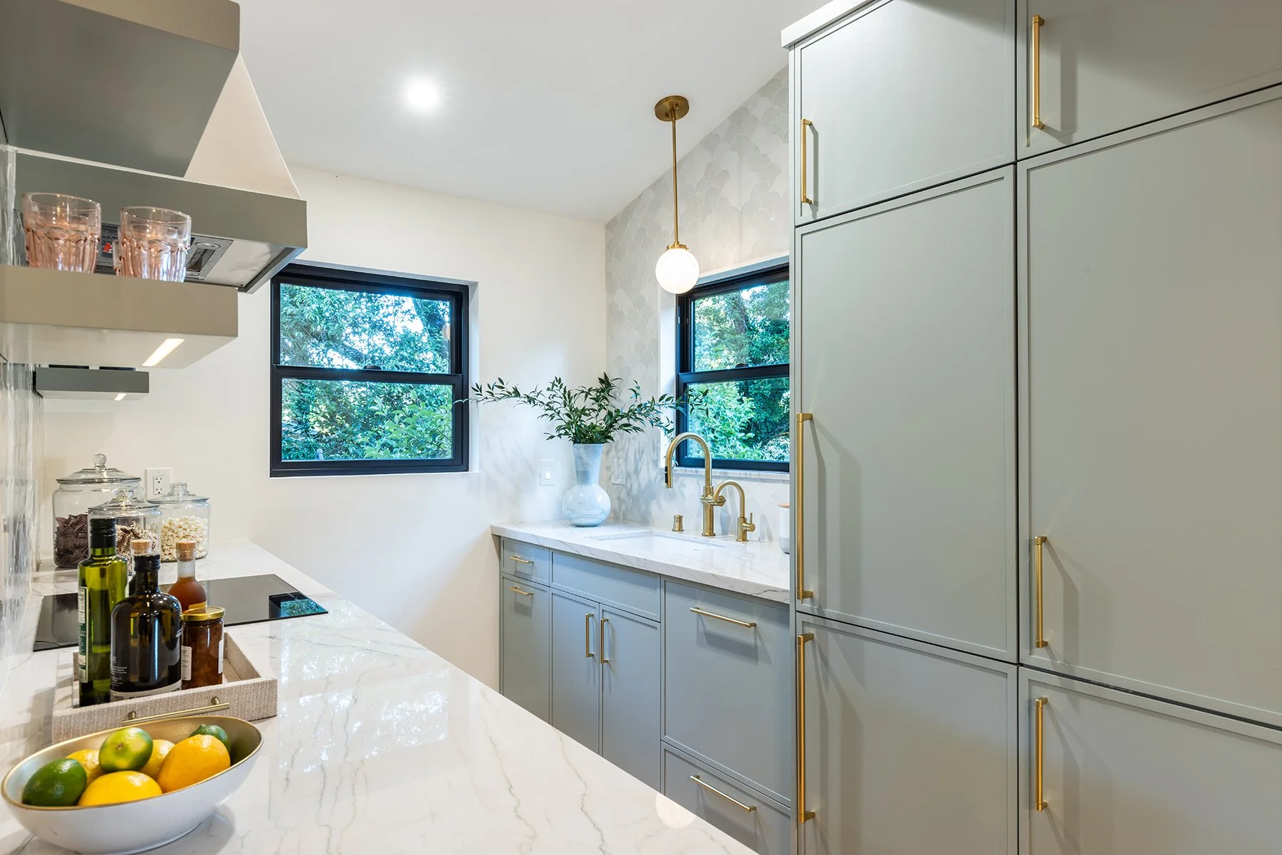Modern kitchen with light gray cabinets, gold handles, marble countertop, and two windows showing greenery outside. There are glass jars with spices, a bowl of lemons and limes, and a potted plant on the counter.