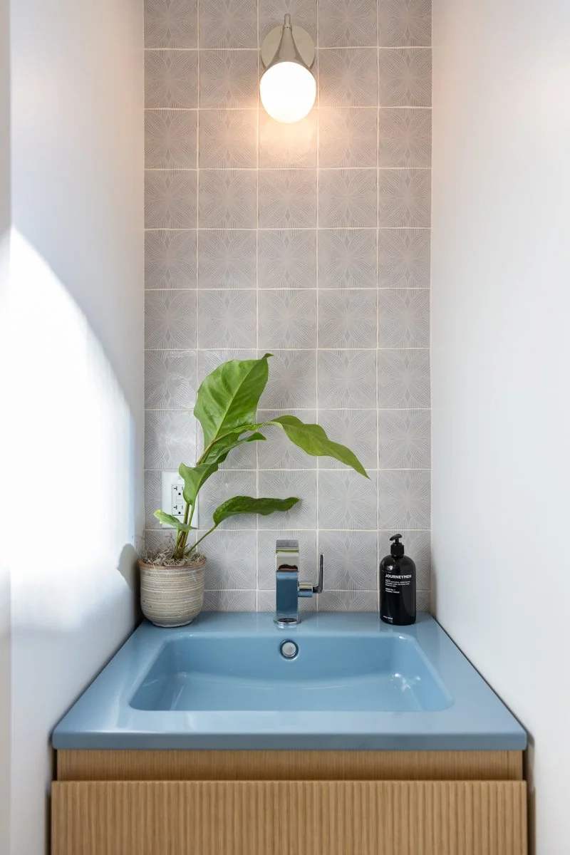 A small bathroom sink with a blue basin, wooden cabinet, a green plant in a pot, a black soap dispenser, and a wall-mounted light fixture above a tiled wall.