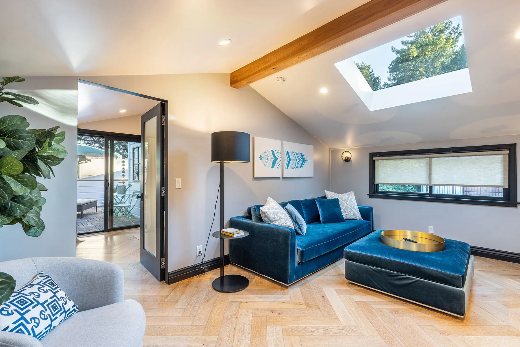 Bright living room with natural light from skylight and window, featuring a blue velvet sofa with pillows, a gold tray table, floor lamp, wall art, and a view of a patio through the door.