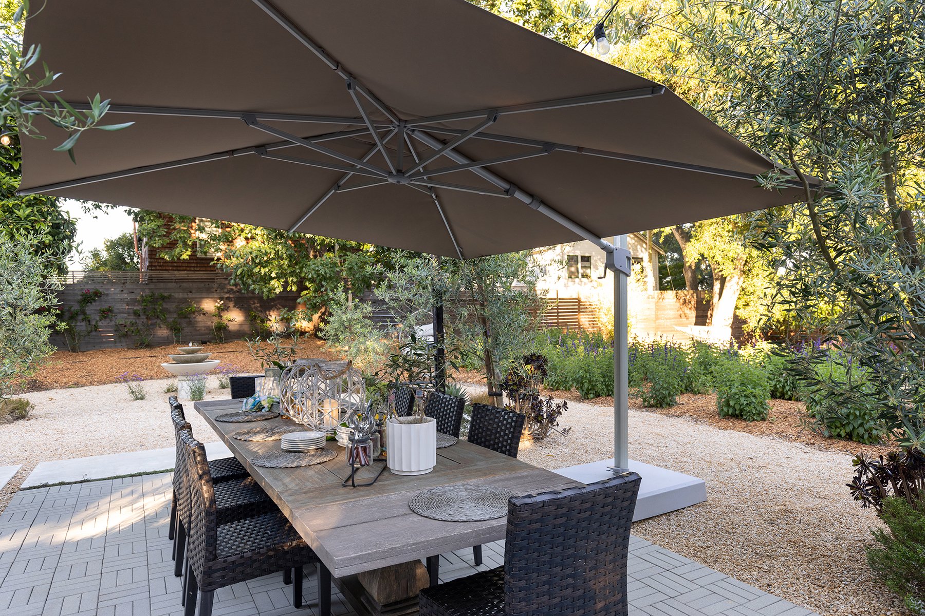 Outdoor dining area with a wooden table, black wicker chairs, and a large beige patio umbrella in a garden setting with trees, plants, and gravel ground.