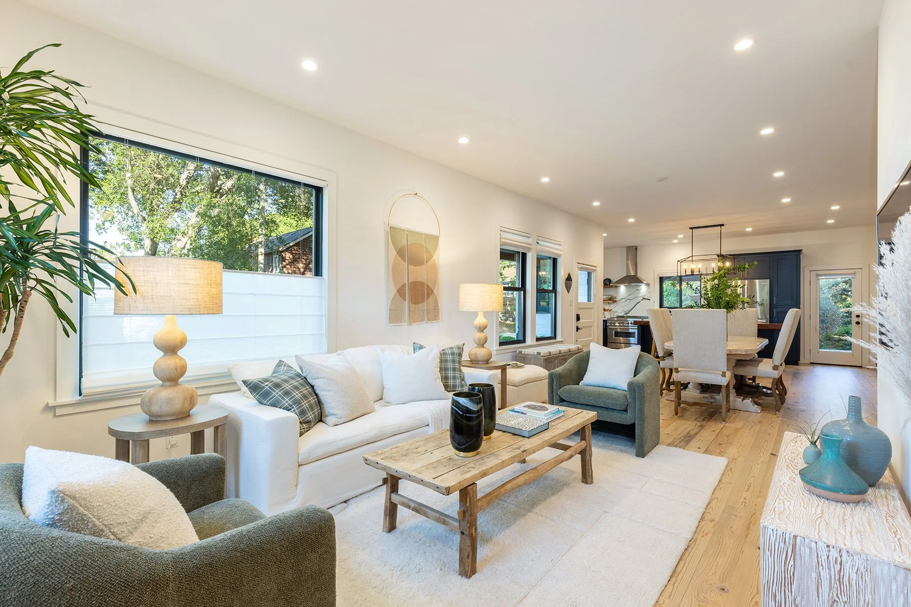 Open-concept living room and dining area with large windows, neutral-colored furniture, wooden accents, and decorative vases.