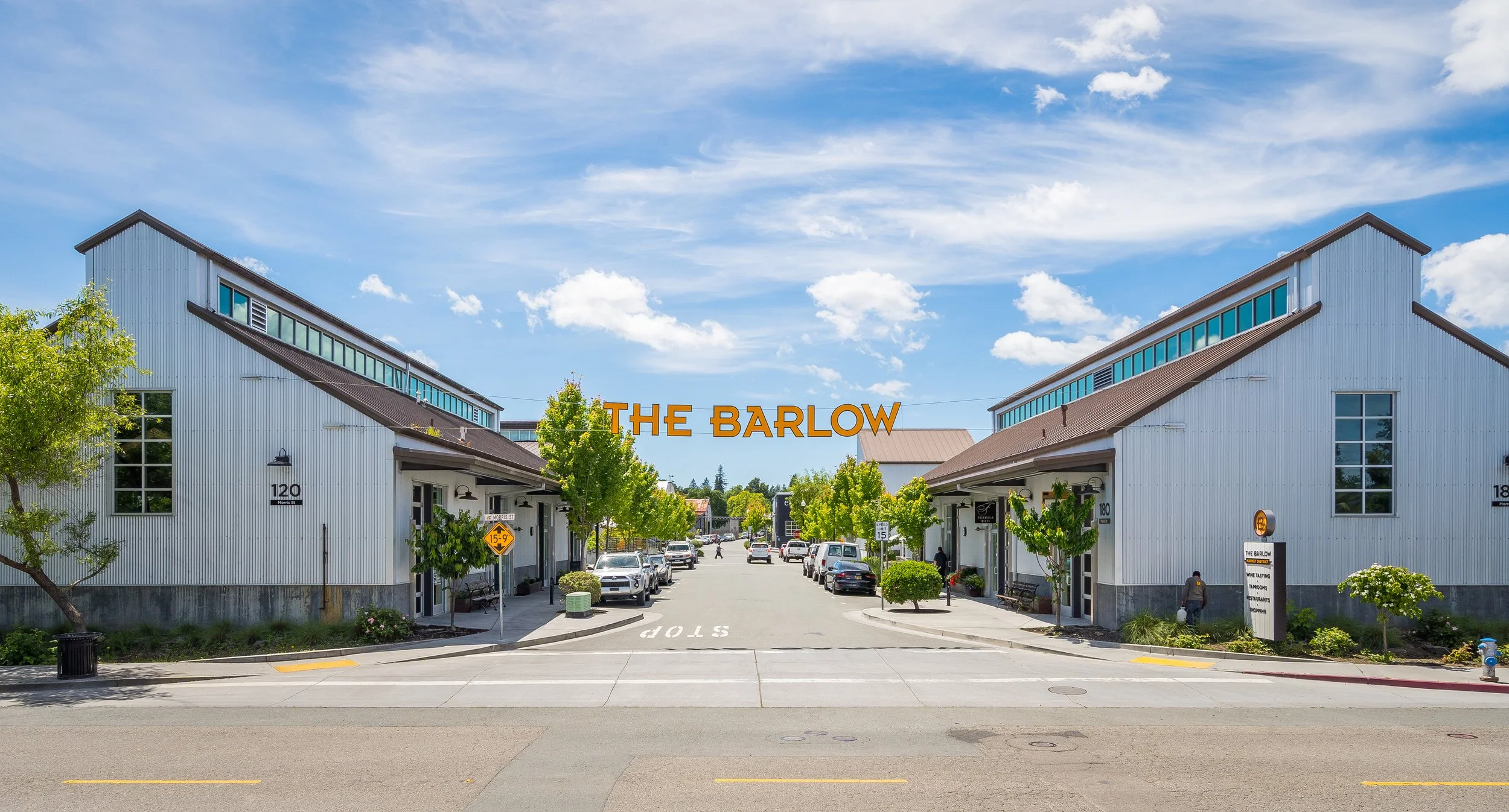 A retail shopping area with two white buildings that have large windows and brown roofs, a sign overhead reading 'The Barlow' in orange letters, trees lining the street, parked cars, and a clear blue sky with some clouds.