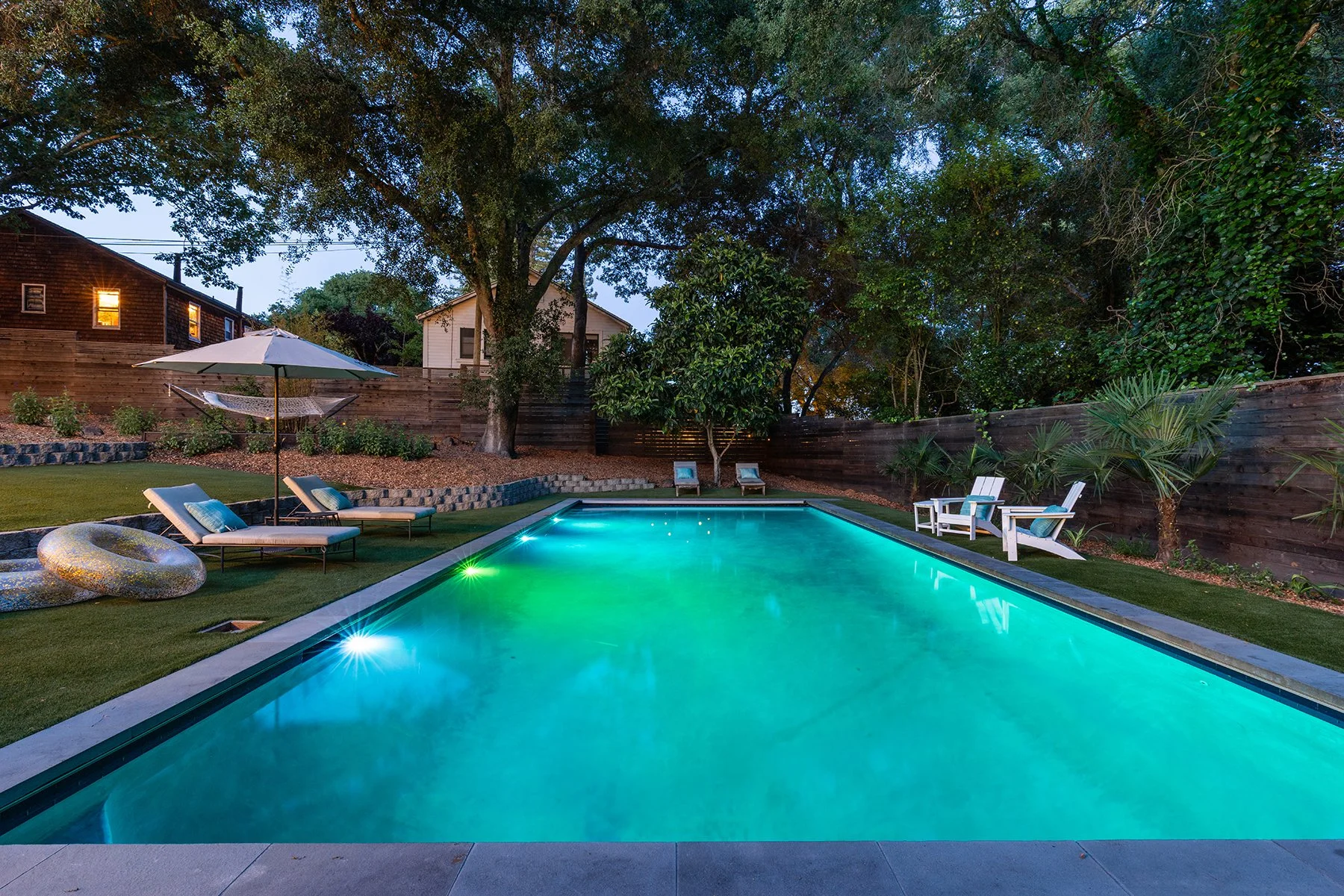 A backyard swimming pool illuminated with green lights at dusk, surrounded by outdoor lounge chairs, a large umbrella, and lush trees and plants.