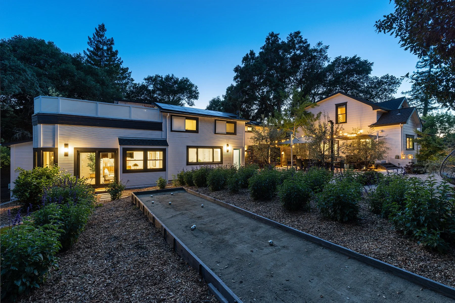 A backyard with a lit house and outdoor patio area, trees, shrubs, and a bocce ball court at twilight.