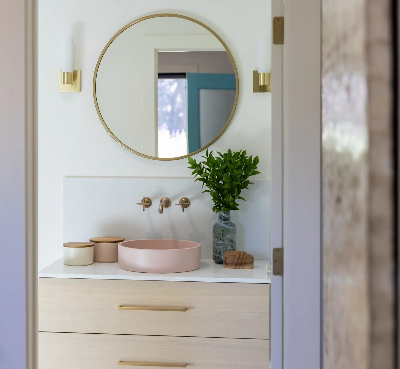 Bathroom vanity area with a round mirror, two wall sconces, a pink sink, and a green plant in a glass vase.