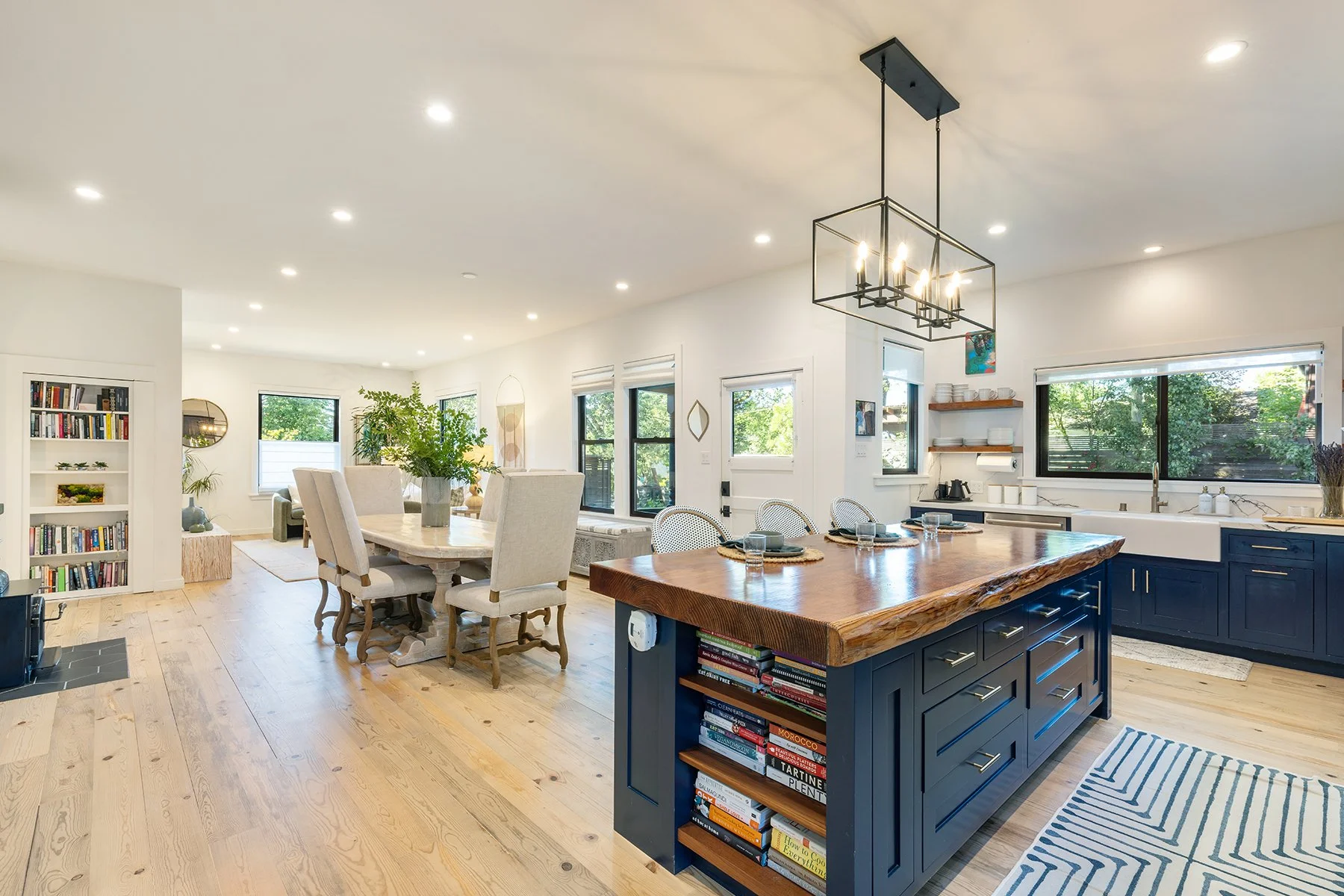 Open-concept kitchen and dining area with light hardwood floors, white walls, large windows, a dark blue kitchen island with bookshelves, a wooden dining table with beige chairs, and a modern chandelier.