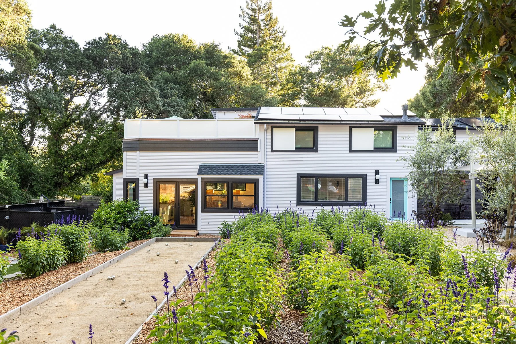 Modern two-story house with black window frames, white siding, and a blue door, surrounded by a lush garden with purple flowers and a sandy bocce court.