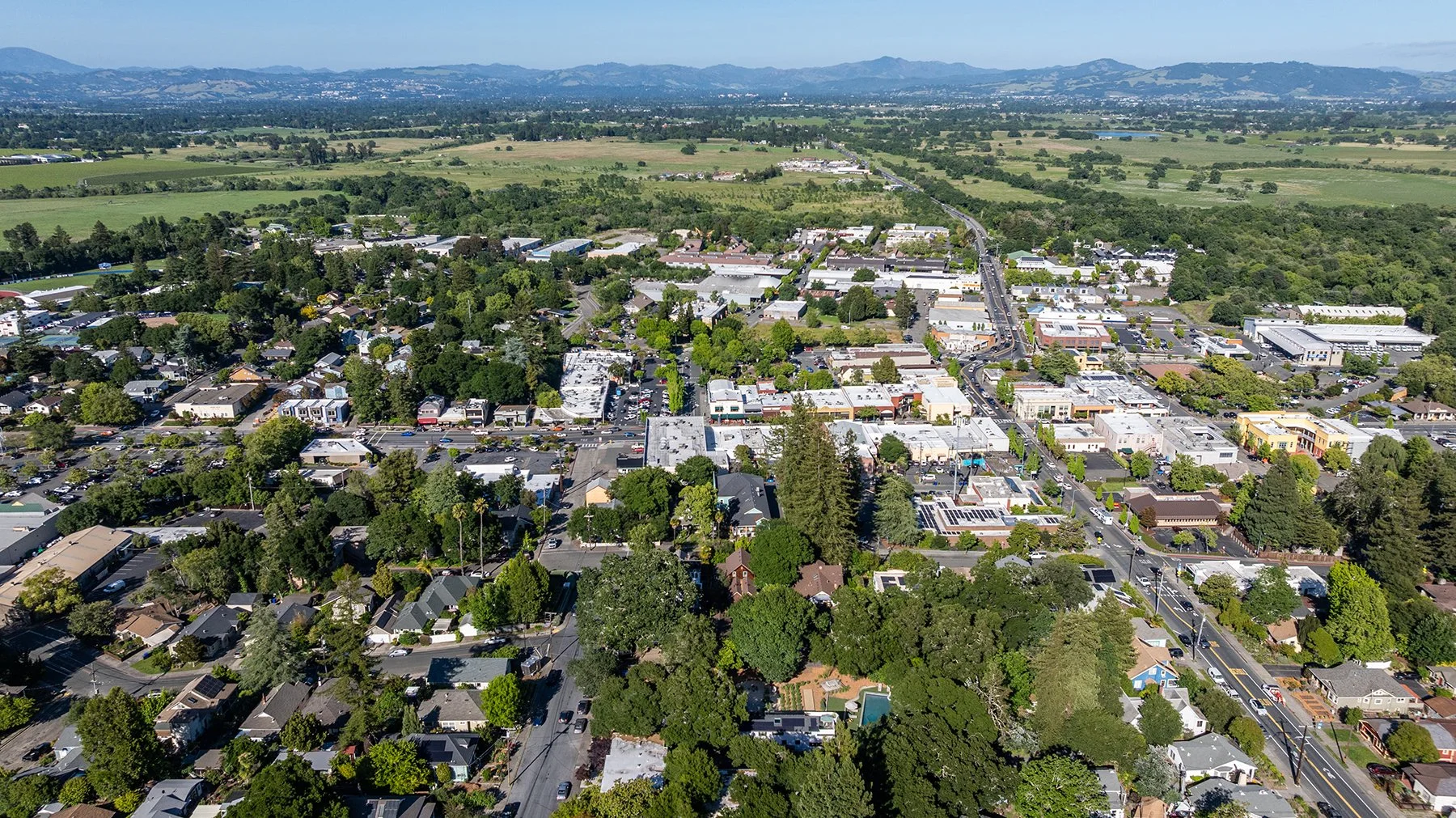 Aerial view of a small town with streets, buildings, and lots of green trees, surrounded by open fields and distant mountains.