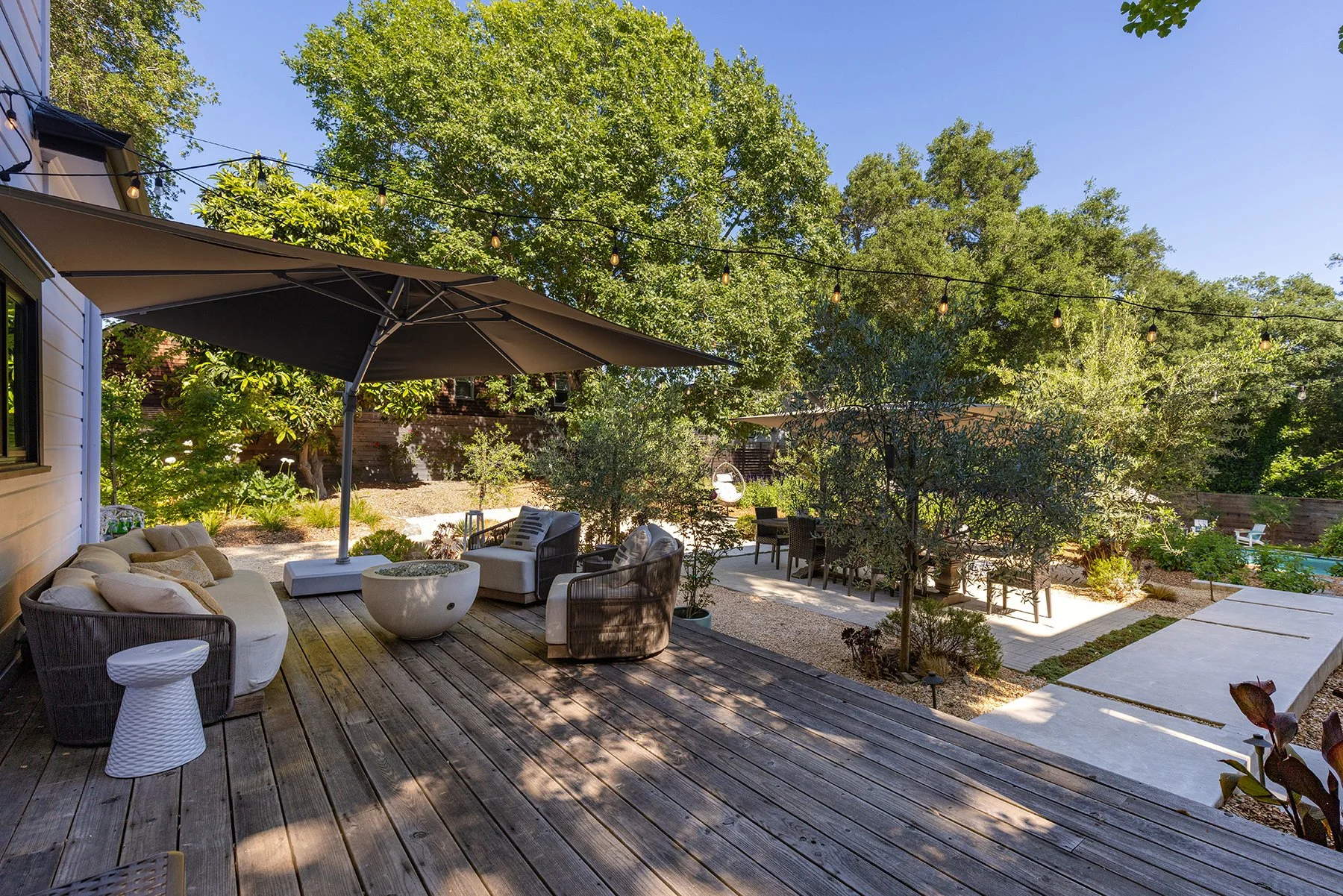 A backyard patio area with wooden decking, outdoor furniture including a white sofa with pillows, armchairs, and a small side table. Large umbrellas provide shade, and string lights are hanging overhead. There are trees, bushes, and a pool in the bac