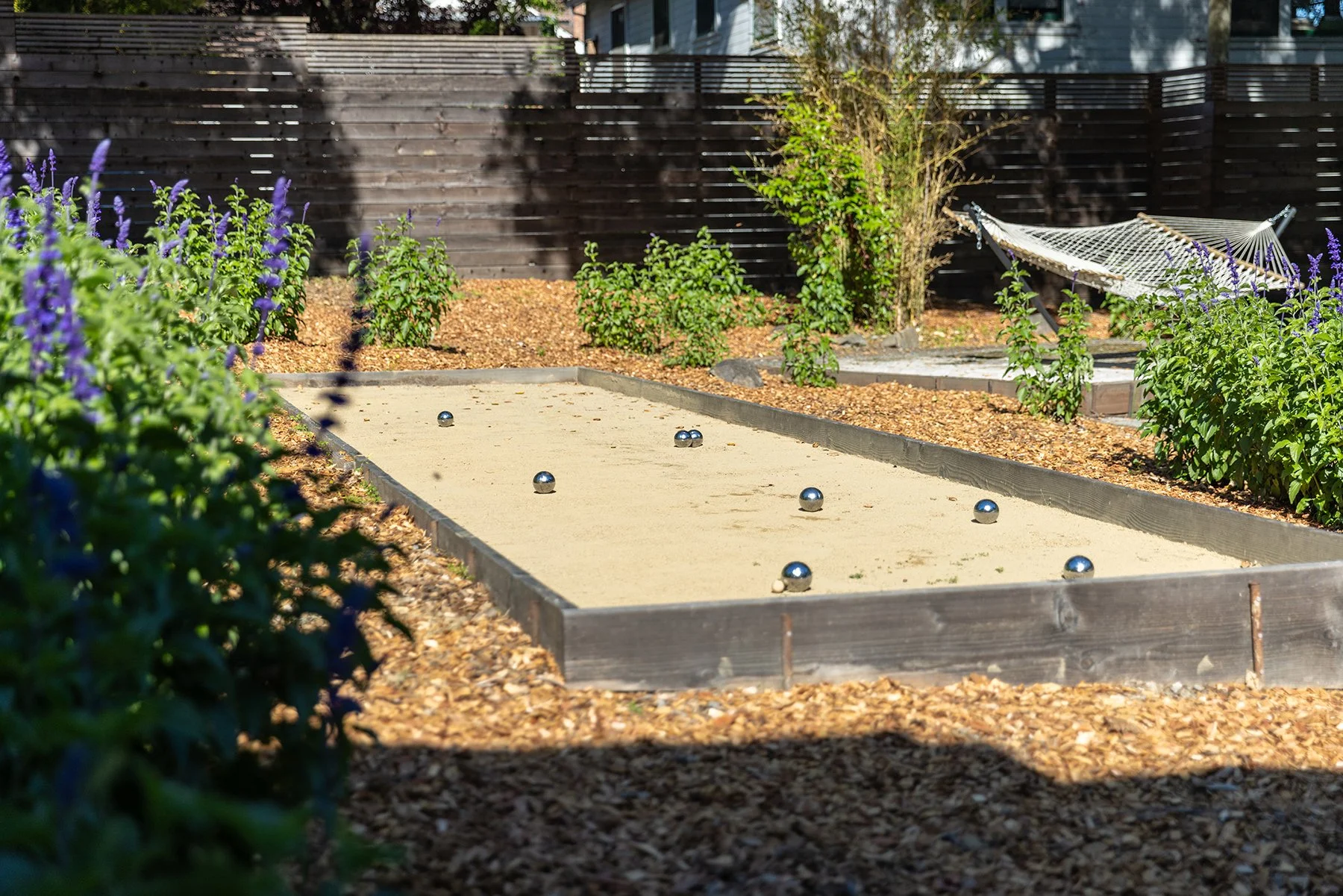 A backyard bocce ball court with several metal balls on the sand playing surface, surrounded by bushes and a wooden fence, with a white hammock in the background.