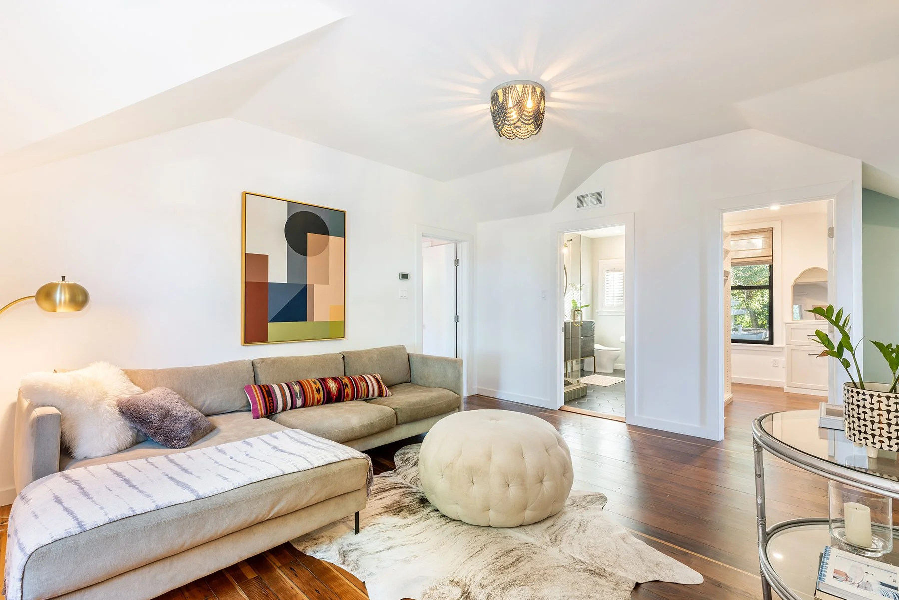 Modern living room with beige sofa, colorful striped pillow, white and purple cushions, gold wall lamp, abstract artwork, white walls, small beige ottoman, cowhide rug, open door to bathroom, and adjacent room with black window and green plant.