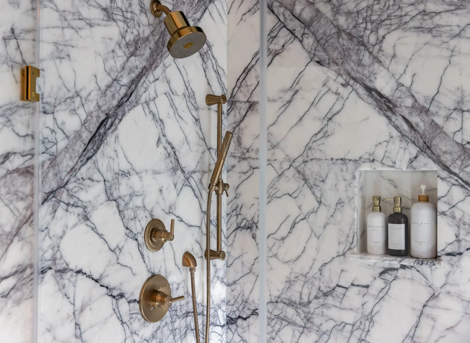 Marble shower with a gold showerhead, handheld shower, and control knobs. A recessed shelf holds white and dark bottles of body wash and lotion.