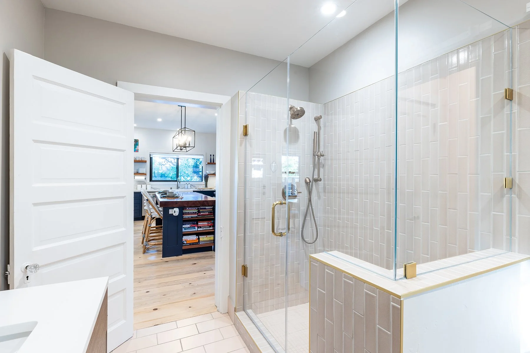 Modern bathroom with a glass-enclosed shower with white tiled walls and gold fixtures, seen from the doorway into a kitchen with a wooden island and open shelving.