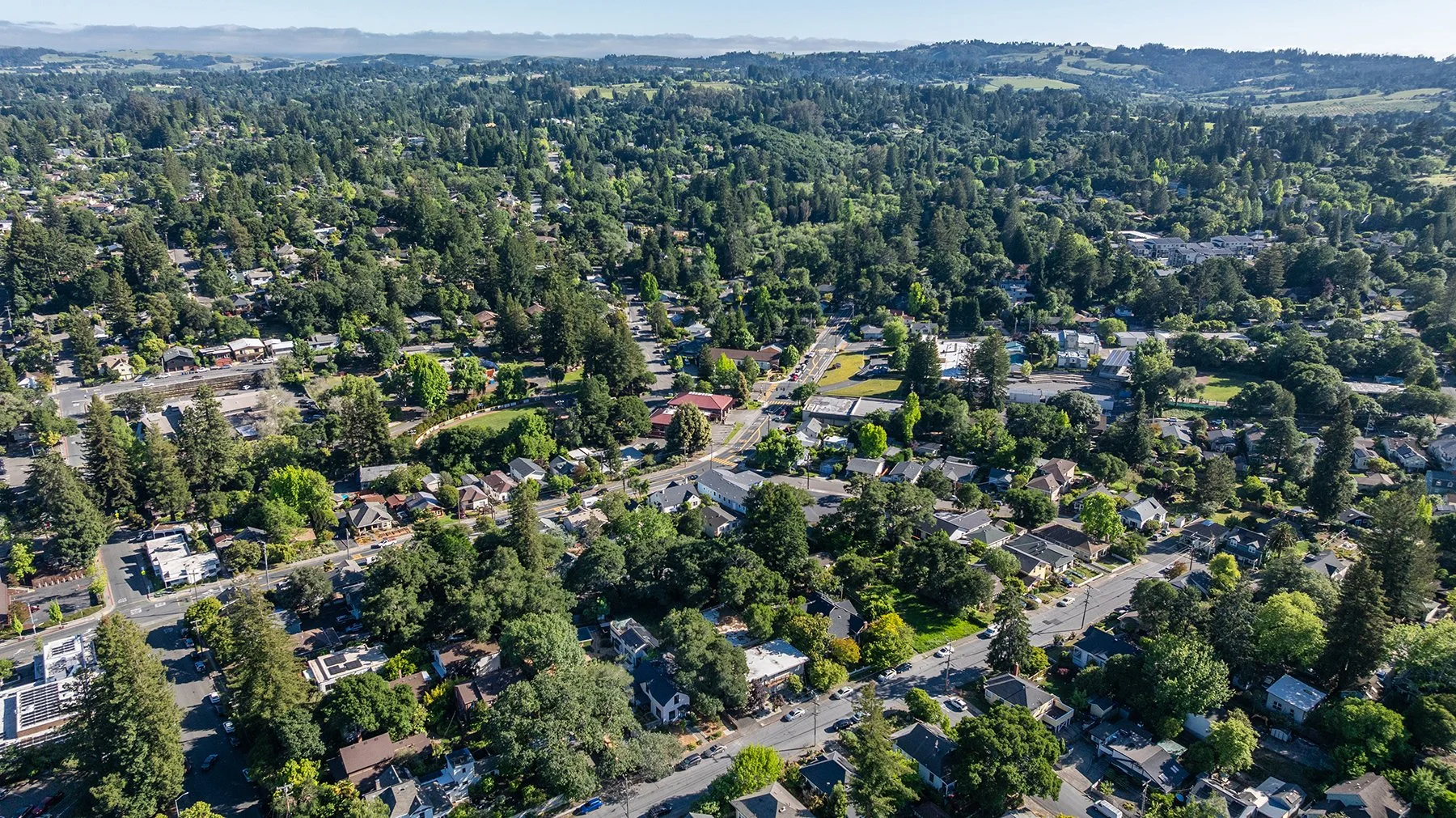 Aerial view of a suburban neighborhood with numerous houses and trees, surrounded by green hills and distant mountains under a partly cloudy sky.