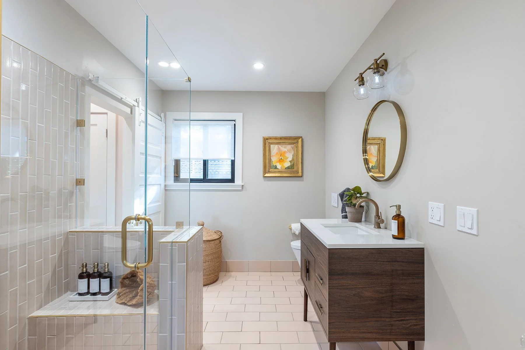 Modern bathroom with tiled shower, vanity with sink and round mirror, window with white blind, framed flower artwork, and decorative items like potted plant and bottles.